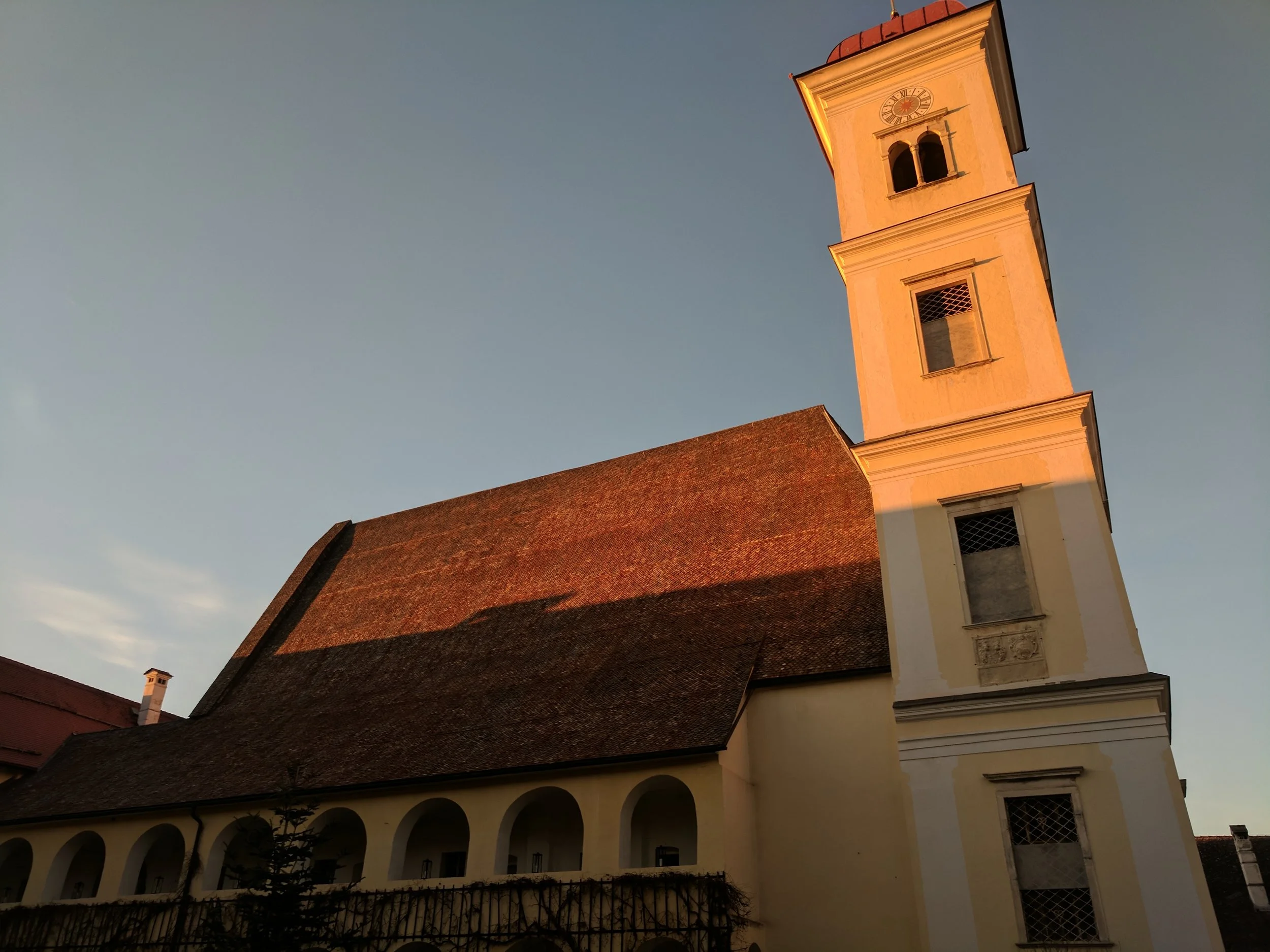 Eine Kirche mit einem hohen gelb-weiß gestrichenen Turm, der eine Uhr hat. Der Kirchturm ist im Sonnenlicht und der Himmel ist blau ohne Wolken.