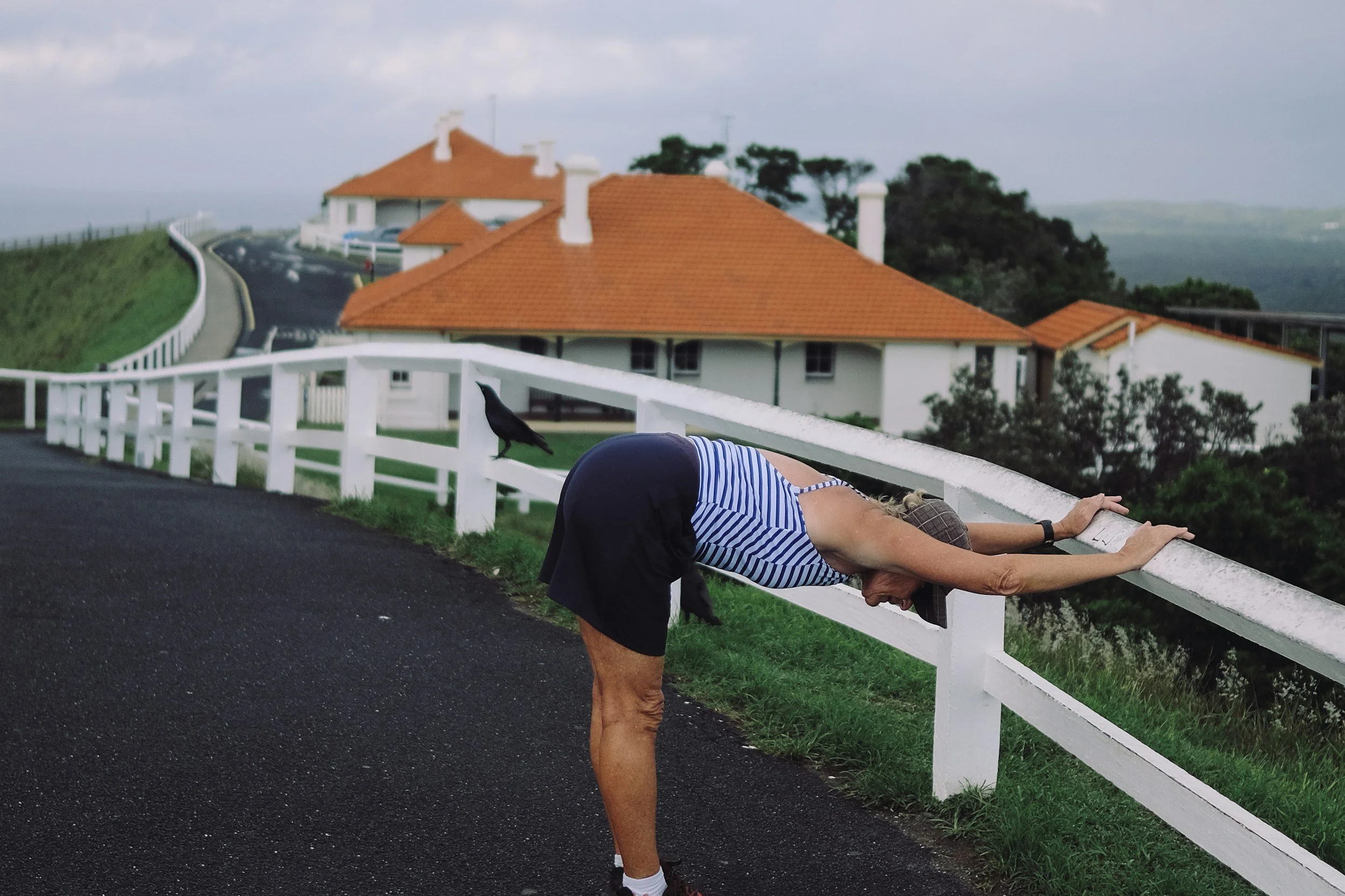 midlife woman wearing tartan cap, blue and white striped top and navy shorts and trainers. Stretching on a white picket fence with houses in the background and a crow on the fence.