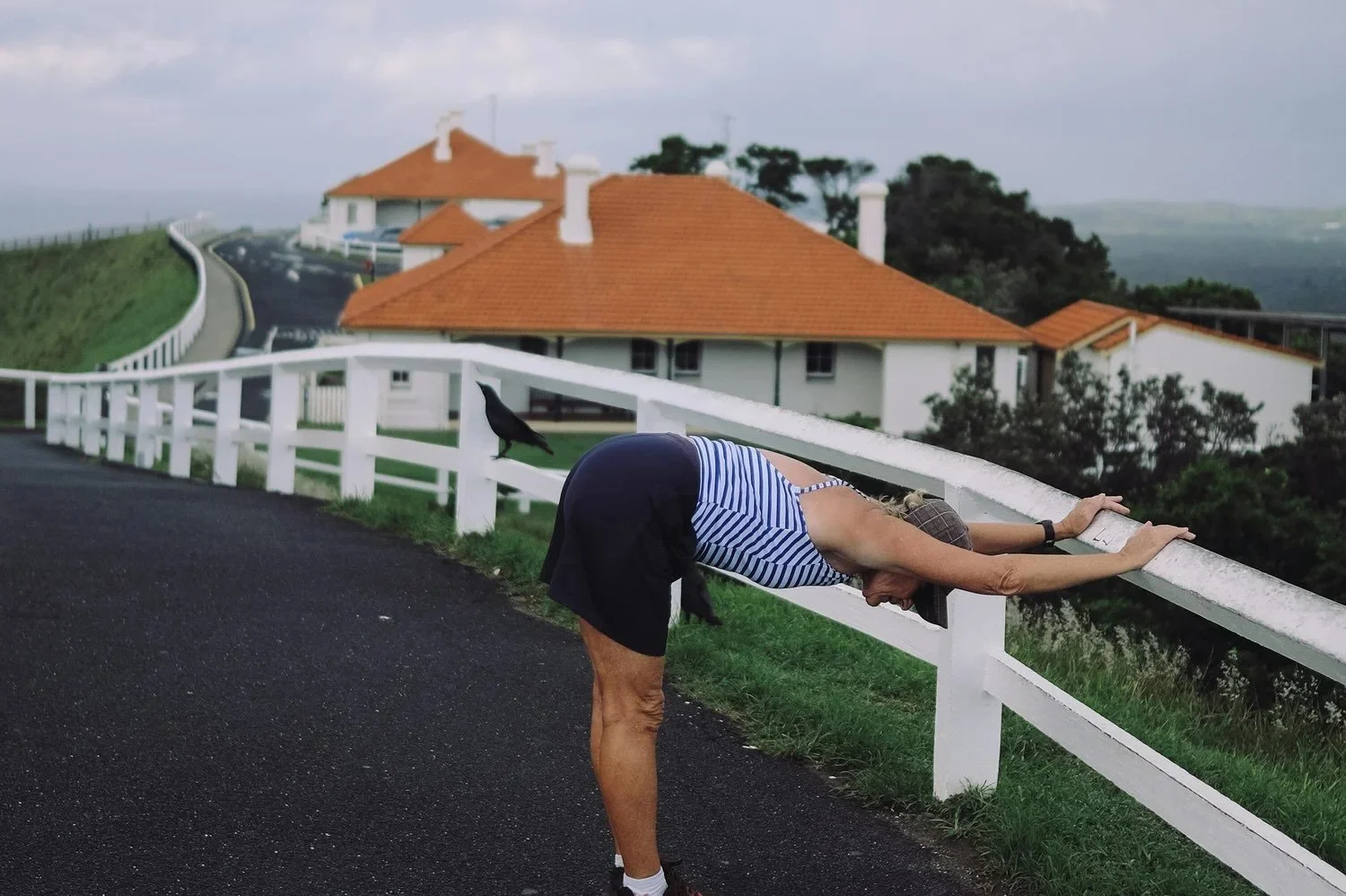 midlife woman wearing tartan cap, blue and white striped top and navy shorts and trainers. Stretching on a white picket fence with houses in the background and a crow on the fence.