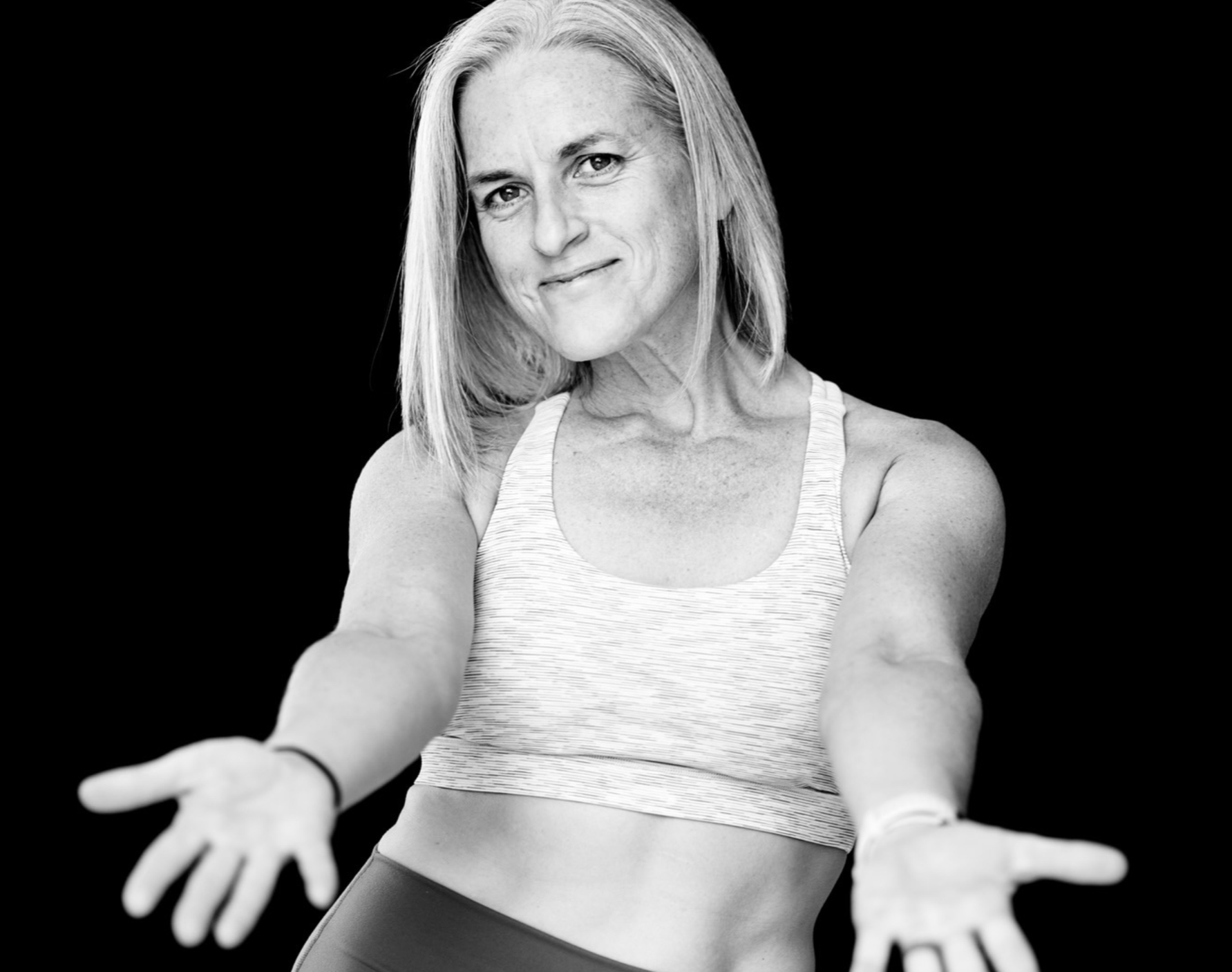 A woman wearing a crop top and short tights smiling with arms out open in an inviting gesture.