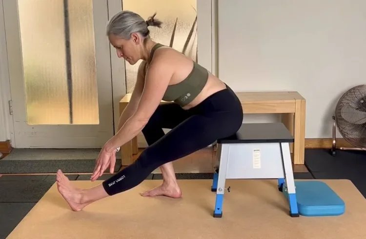 a woman sitting on a stool in a home gym doing a hamstring mobility stretch