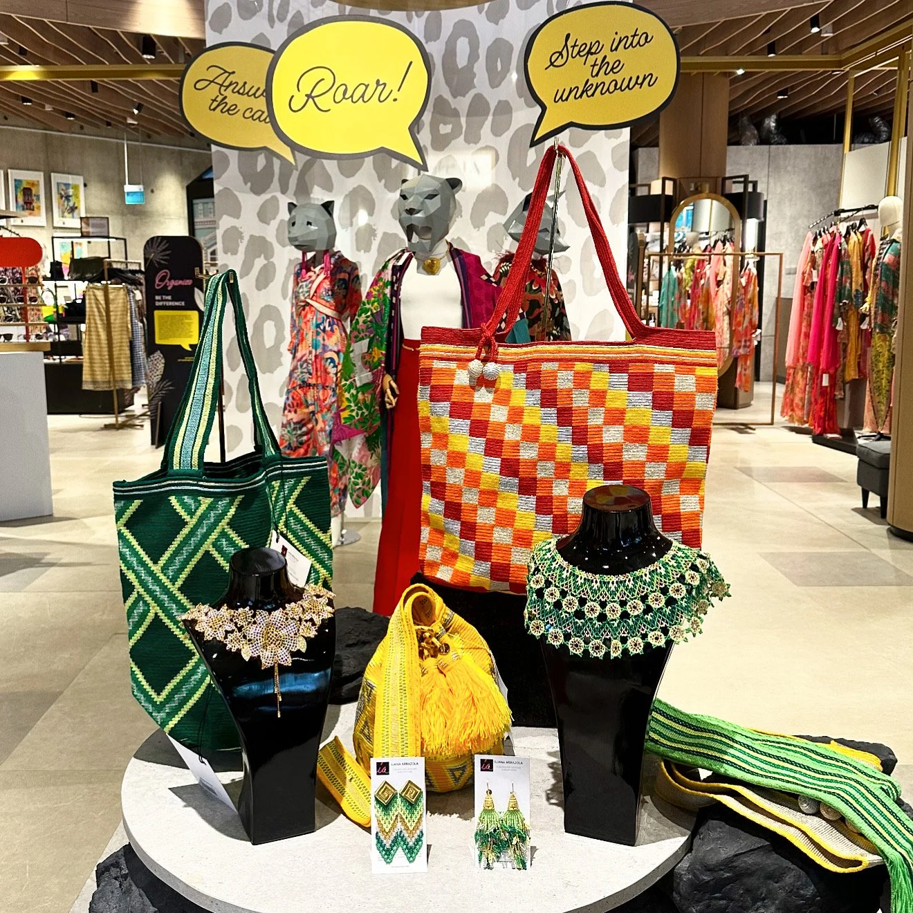 Display of colorful woven handbags, jewelry, and accessories in a retail store with mannequins dressed in vibrant clothing and motivational speech bubbles overhead.