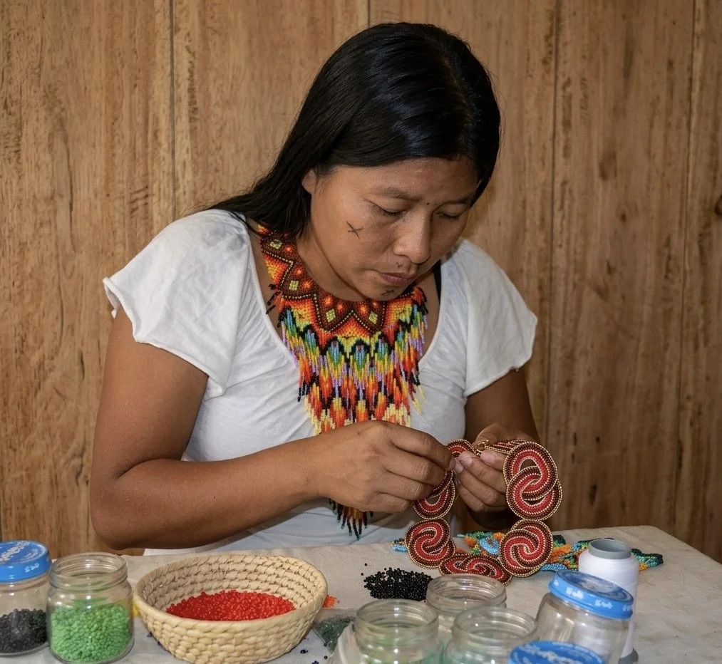 Woman working on beaded jewelry, surrounded by jars of beads, seated at a table with a wood-paneled background.