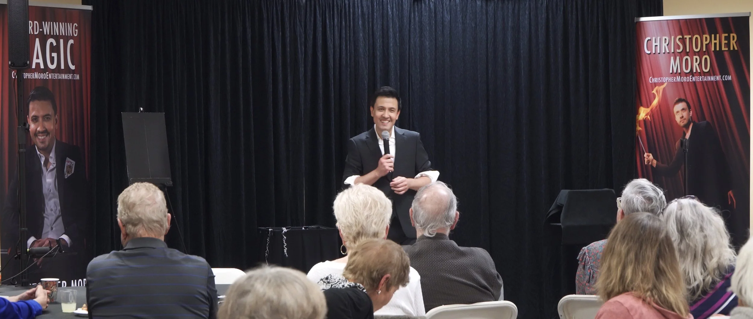 Christopher Moro performing a stand-up stage magic show for a seated audience in Southern California, with branded banners visible on stage