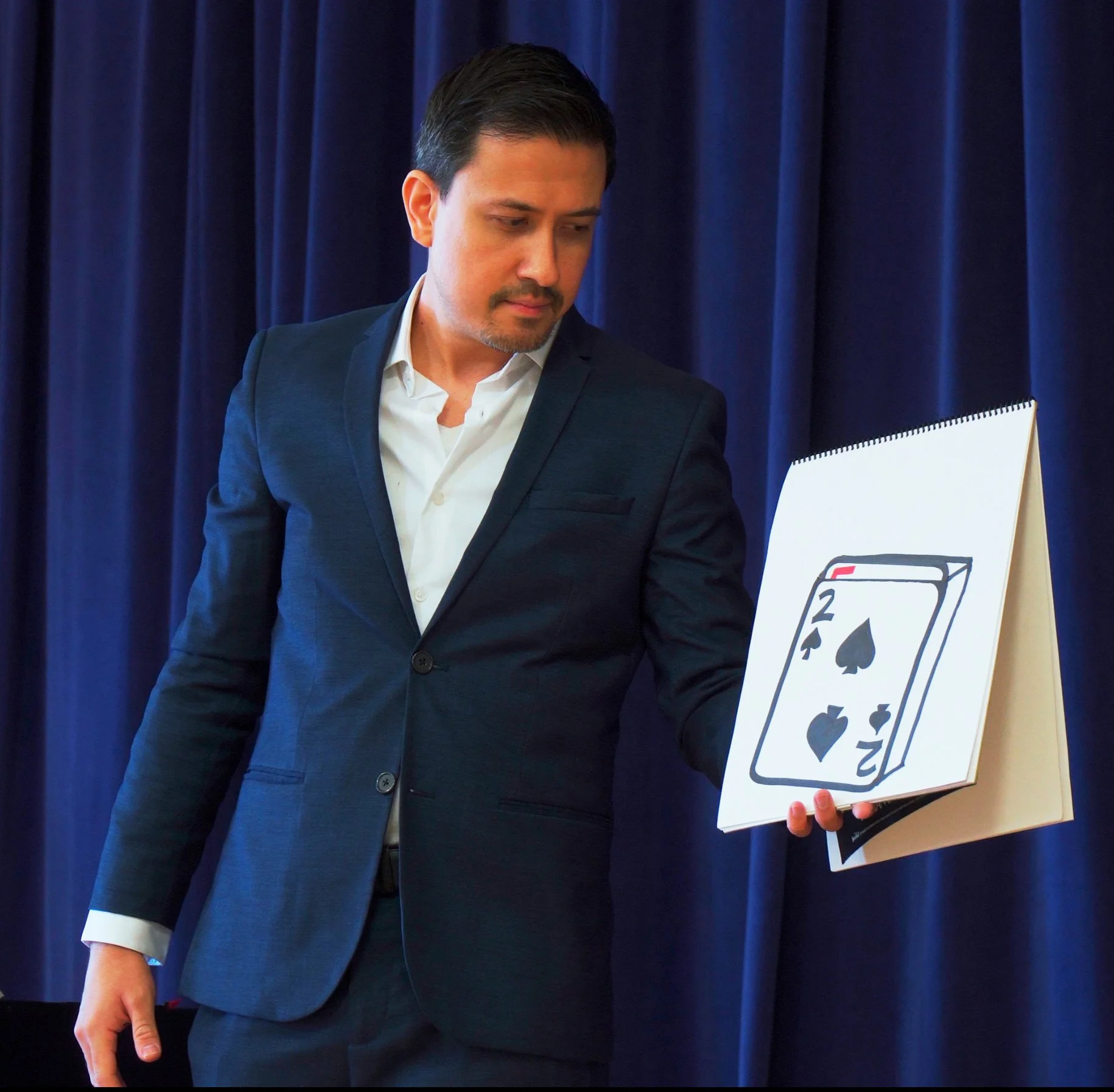 Christopher Moro holding a large sketch pad with a hand-drawn playing card during a stage magic performance in Los Angeles
