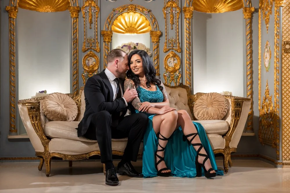 Couple seated together on an ornate sofa, wearing a turquoise dress and dark suit.