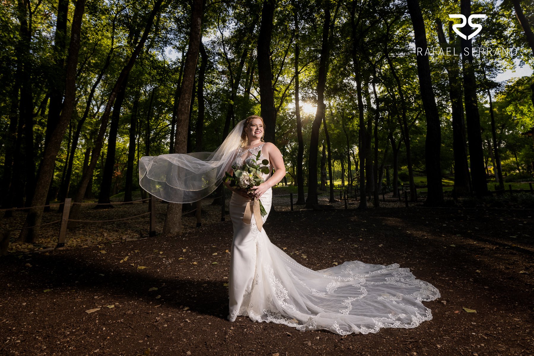 Bride wearing a fitted wedding dress and flowing veil in an outdoor wooded setting.