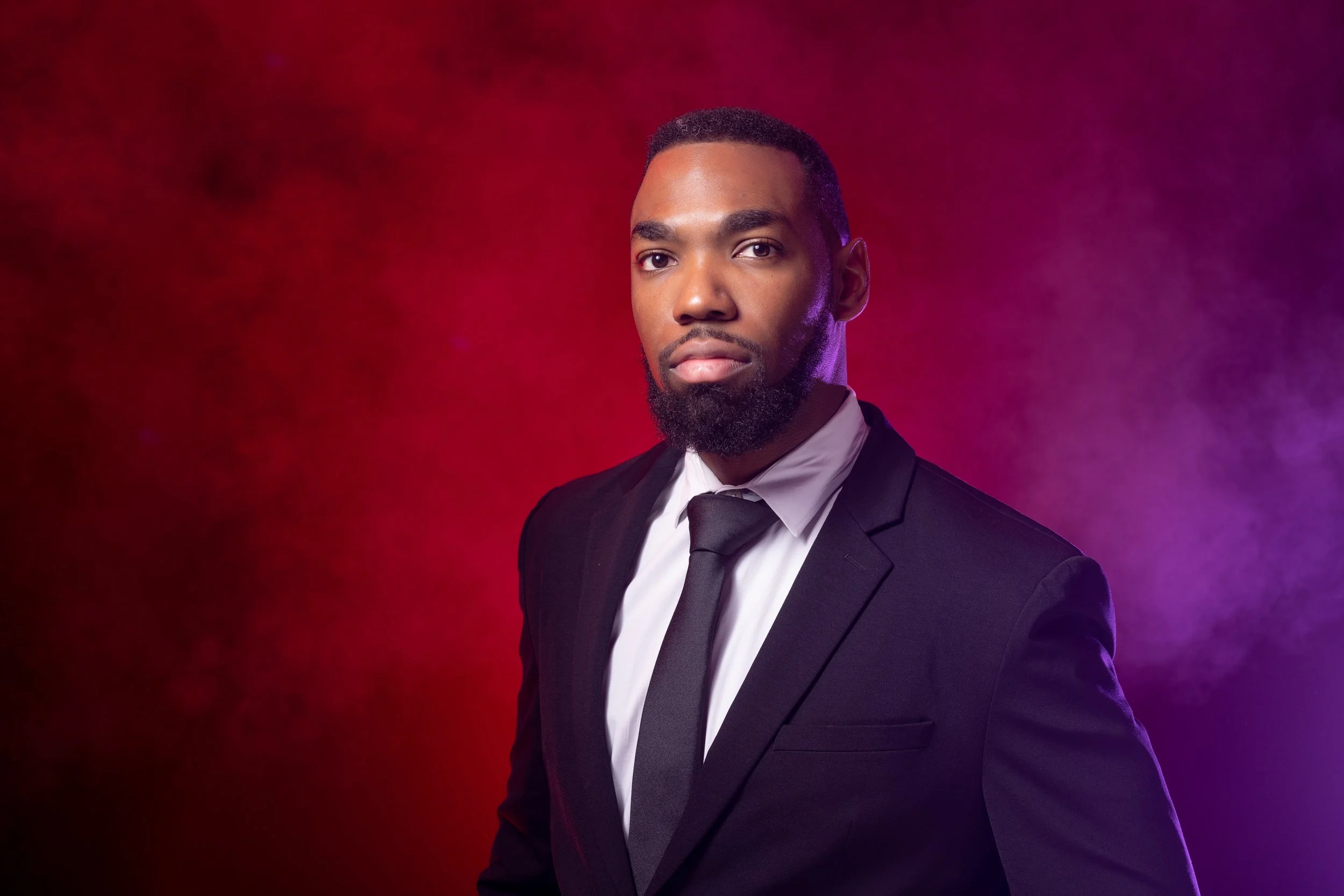Professional headshot of a man wearing a black suit and tie against a dramatic red and purple studio background.