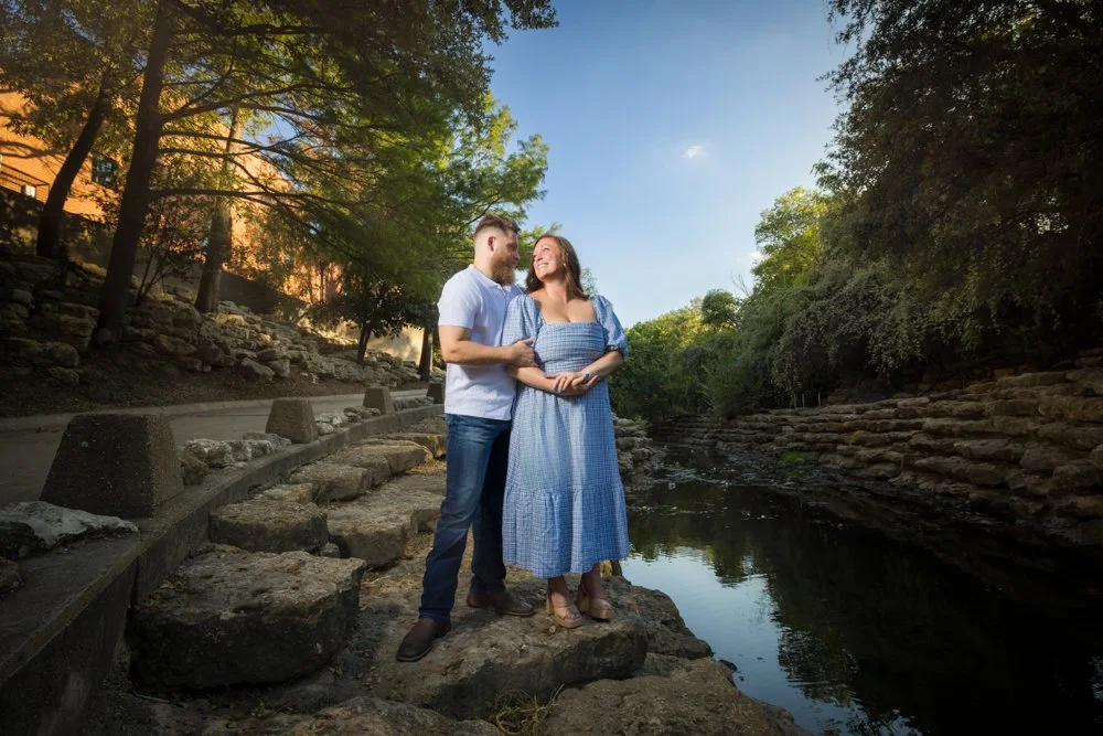 Lacey and Trey facing each other on rocks beside a tree-lined waterway.