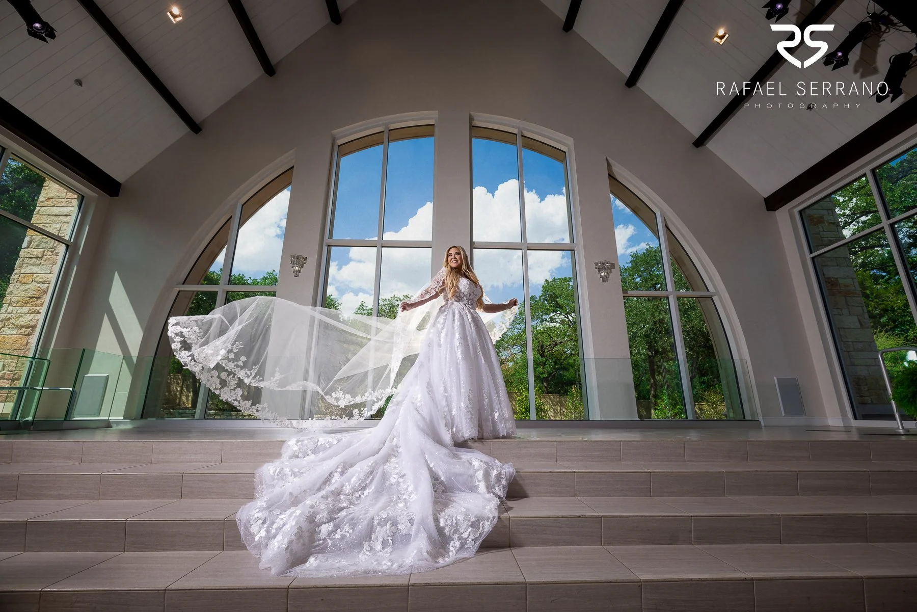 Bride wearing a detailed wedding gown and flowing veil inside an elegant wedding venue.