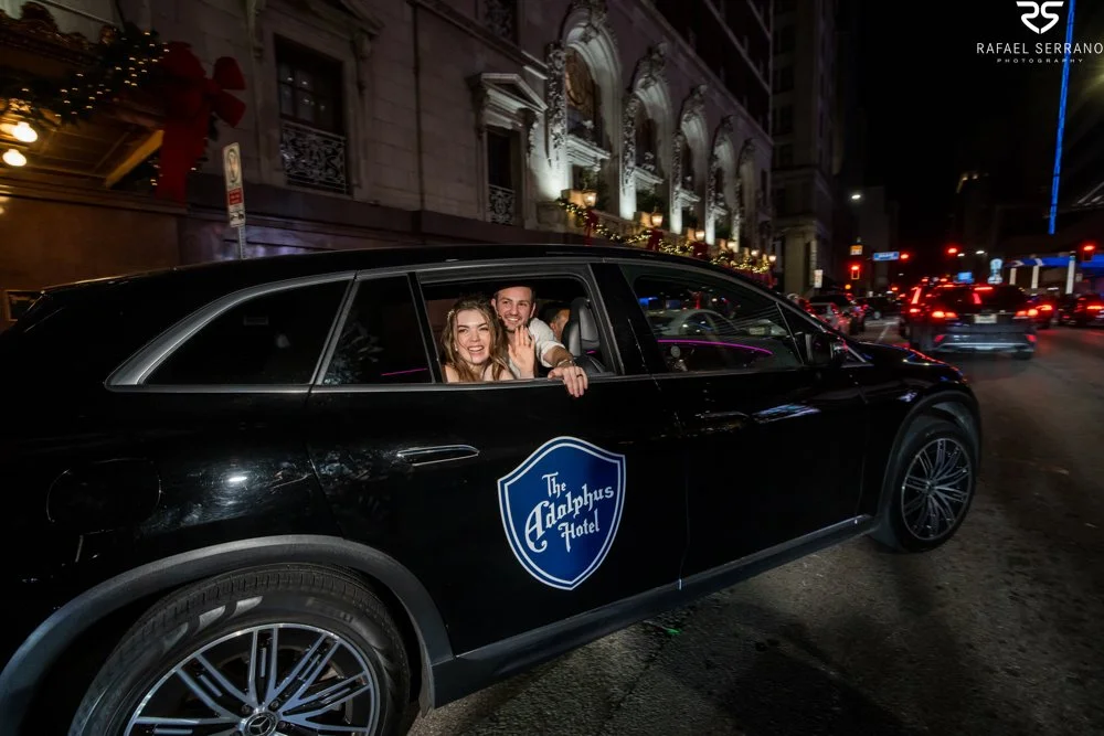 McKenna and Chris waving from an Adolphus-branded vehicle outside the hotel at night.