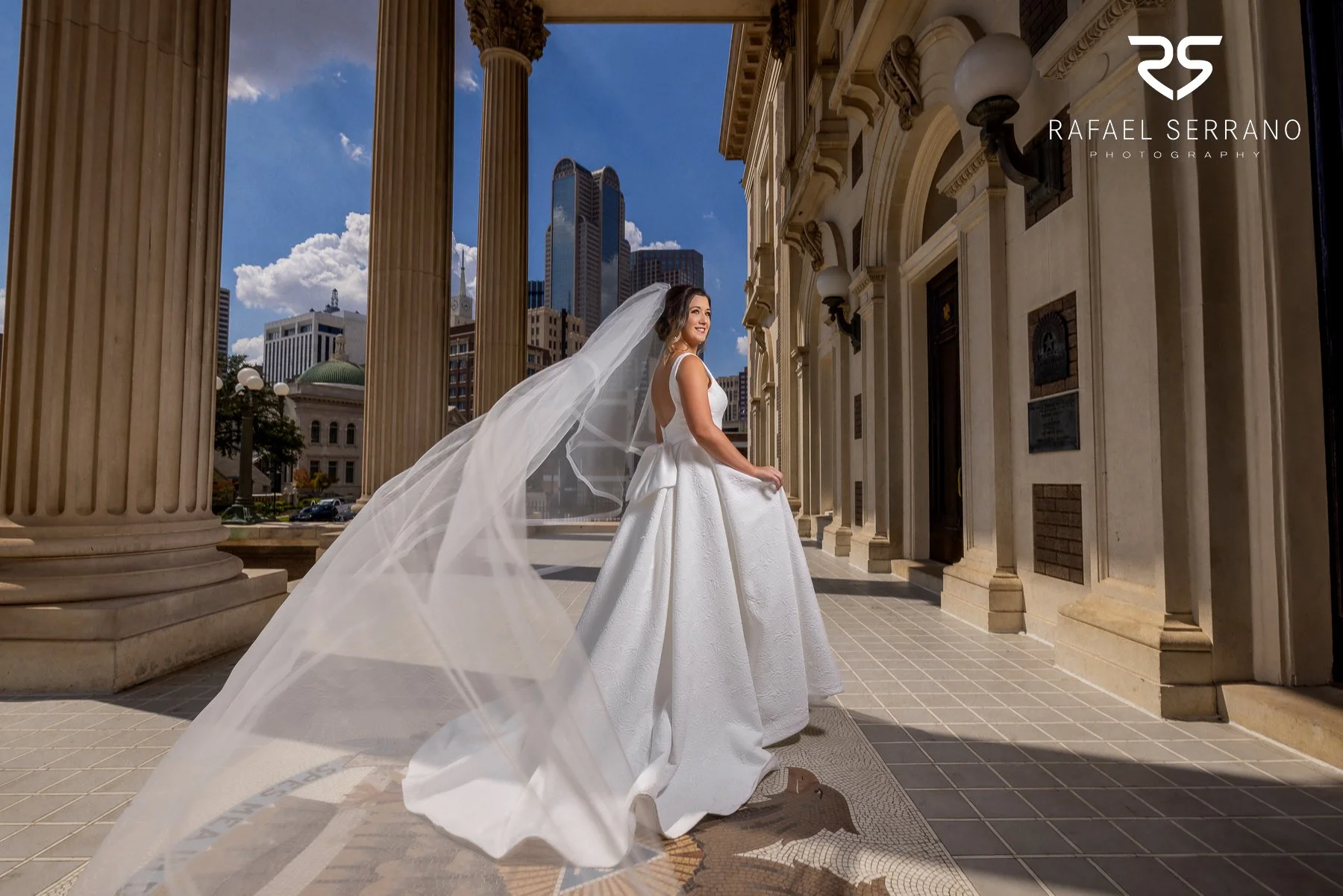 Bride wearing a wedding dress and flowing veil in downtown Dallas with the city skyline behind her.