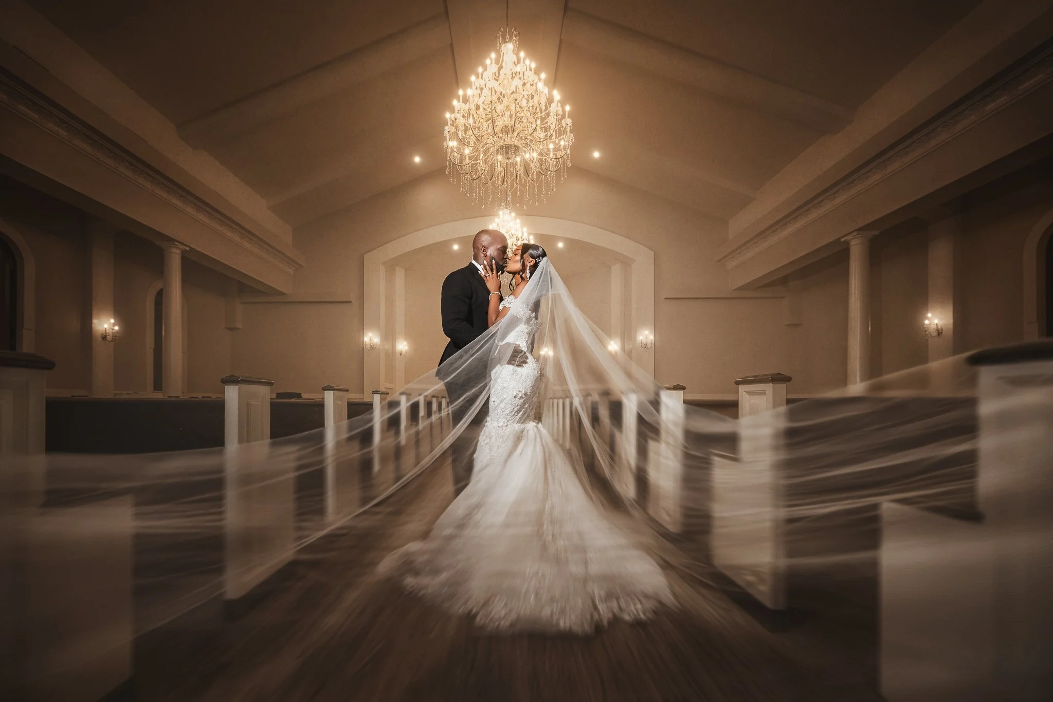 Bride and groom photographed beneath a chandelier inside an elegant church with dramatic veil movement.