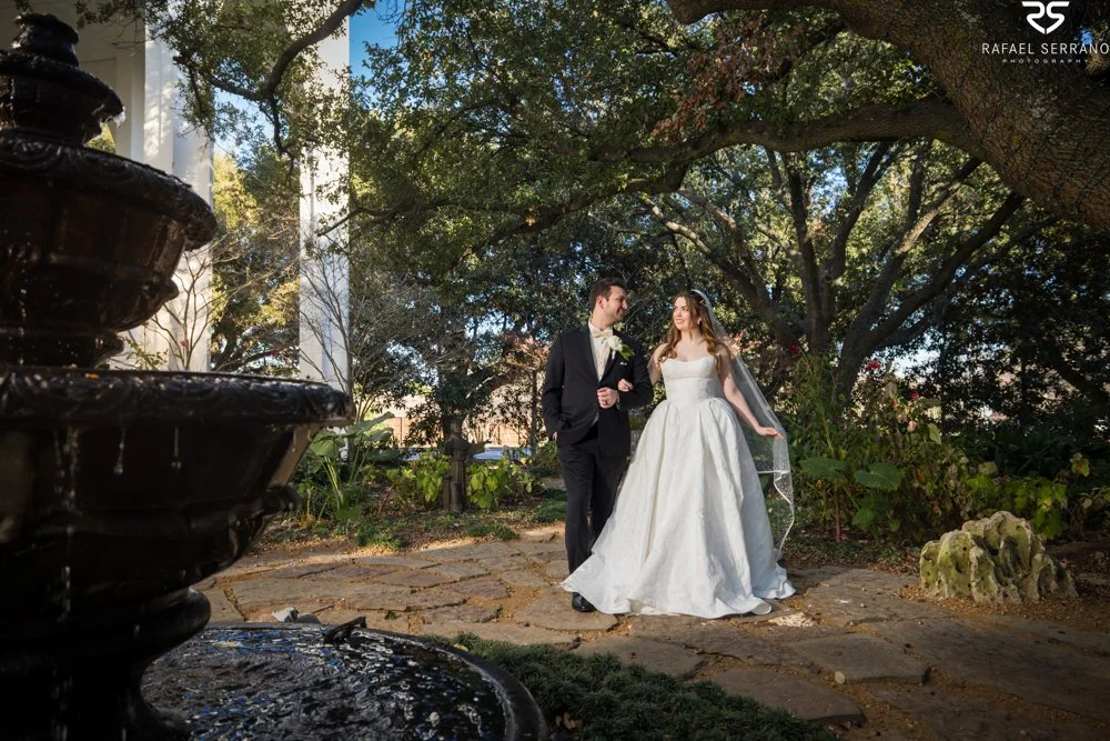 McKenna and Chris standing together beside a fountain beneath spreading trees.