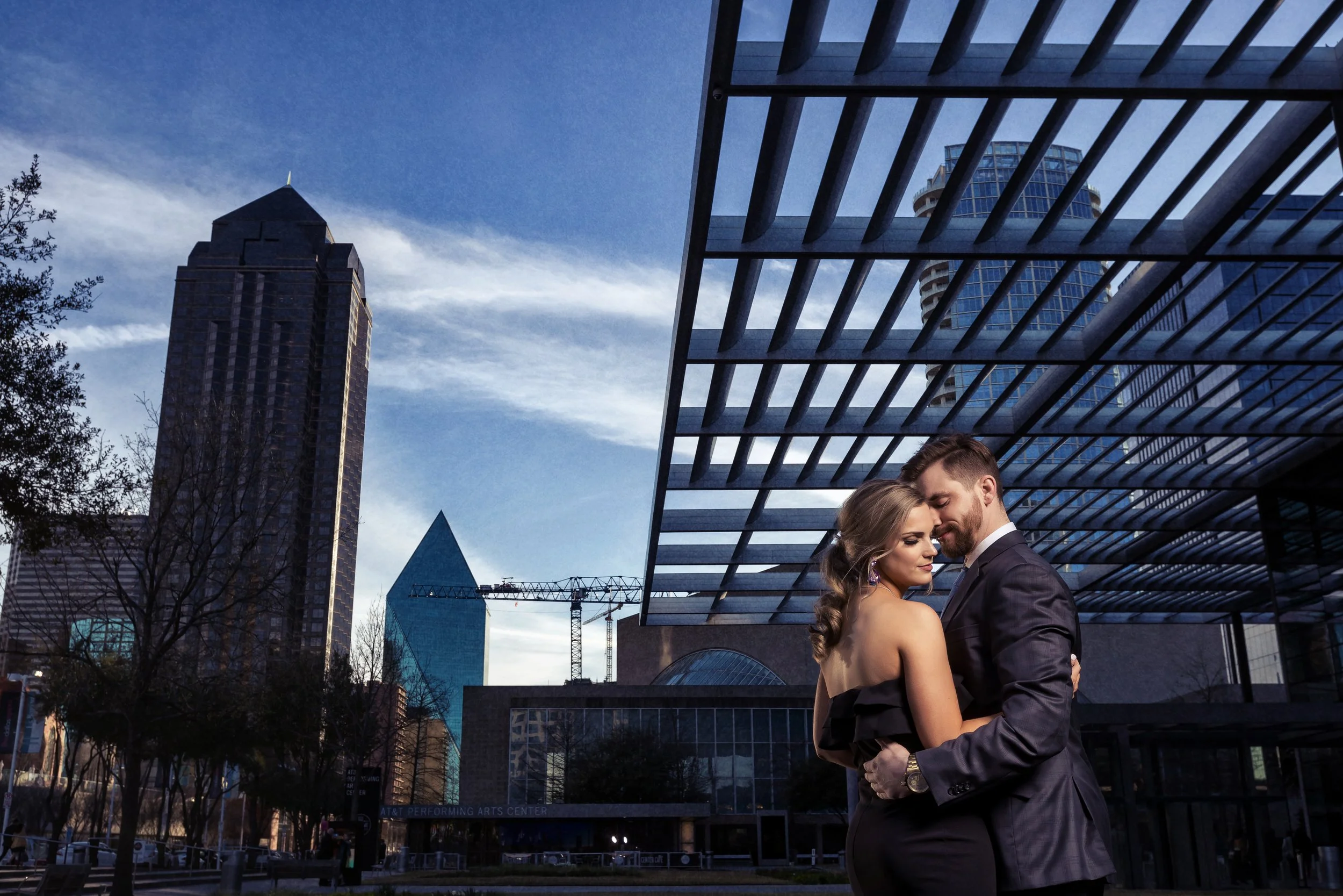 Couple embracing during an engagement photography session in downtown Dallas with the city skyline in the background.