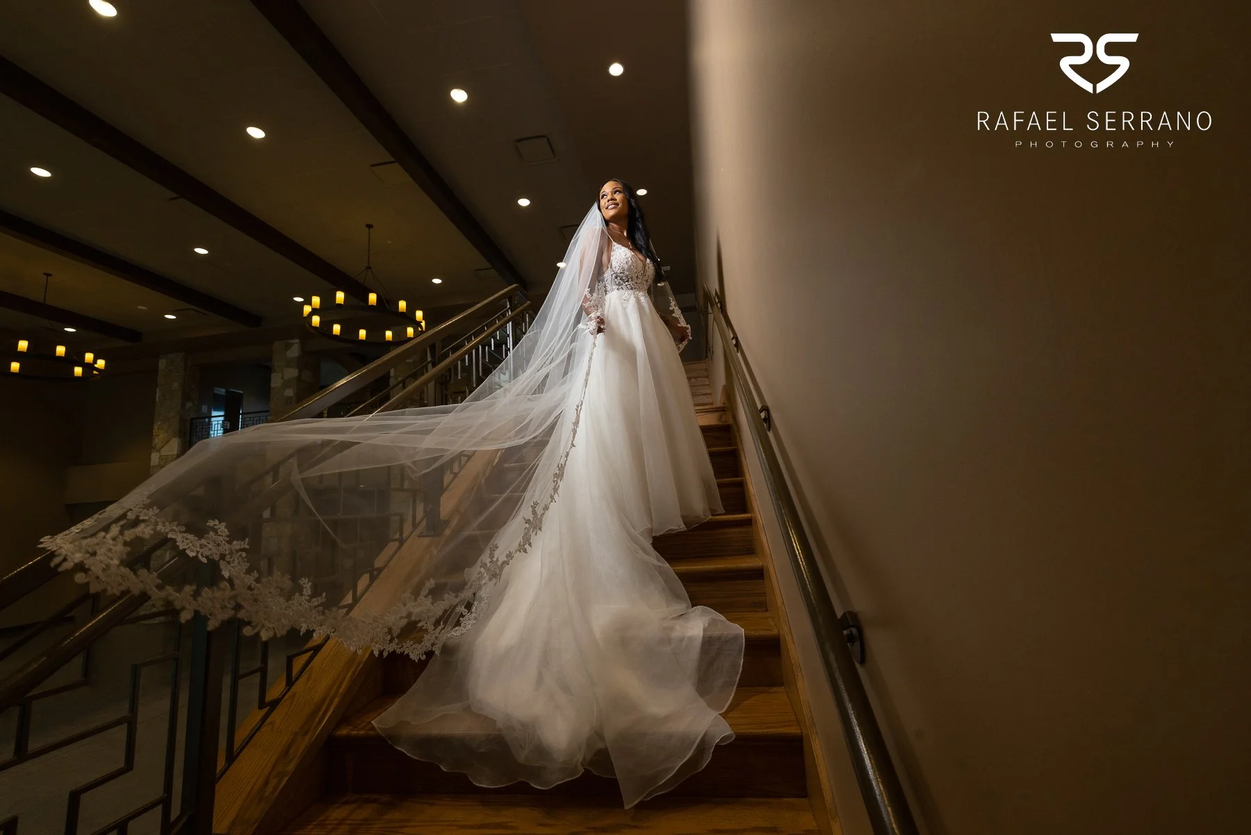 Bride wearing a wedding dress and long flowing veil on an indoor staircase.