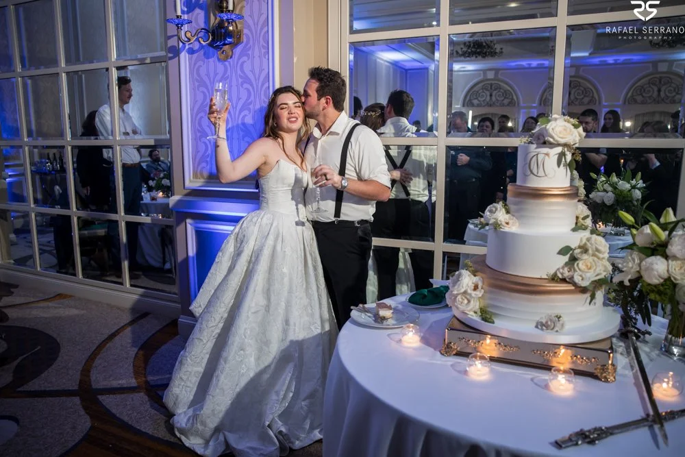 McKenna raising a glass as Chris kisses her beside their white tiered wedding cake.