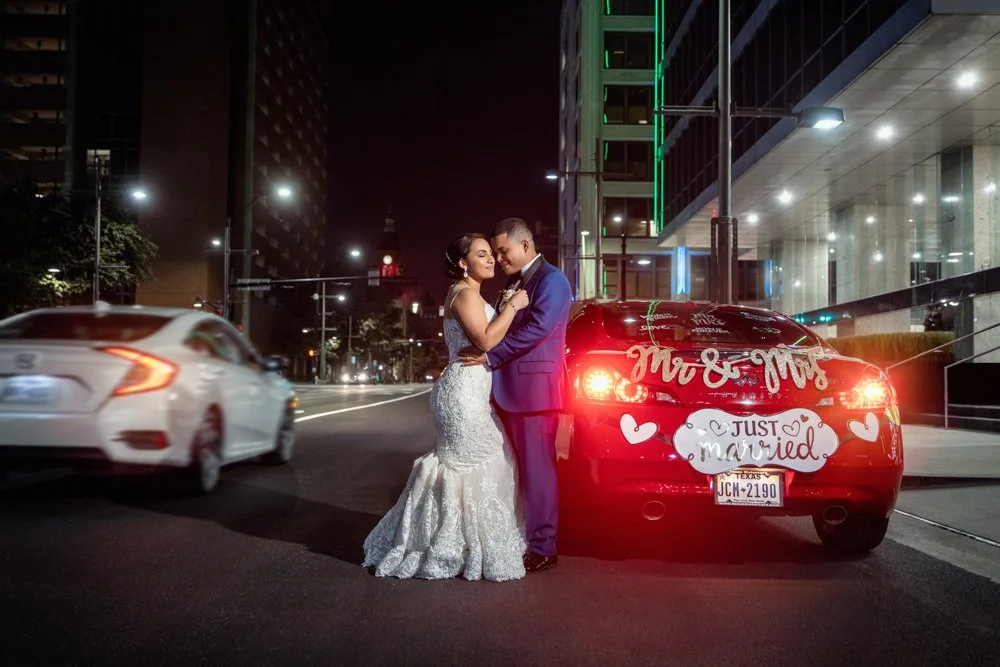 Newlywed couple posing beside a decorated getaway car on a downtown Dallas street at night.