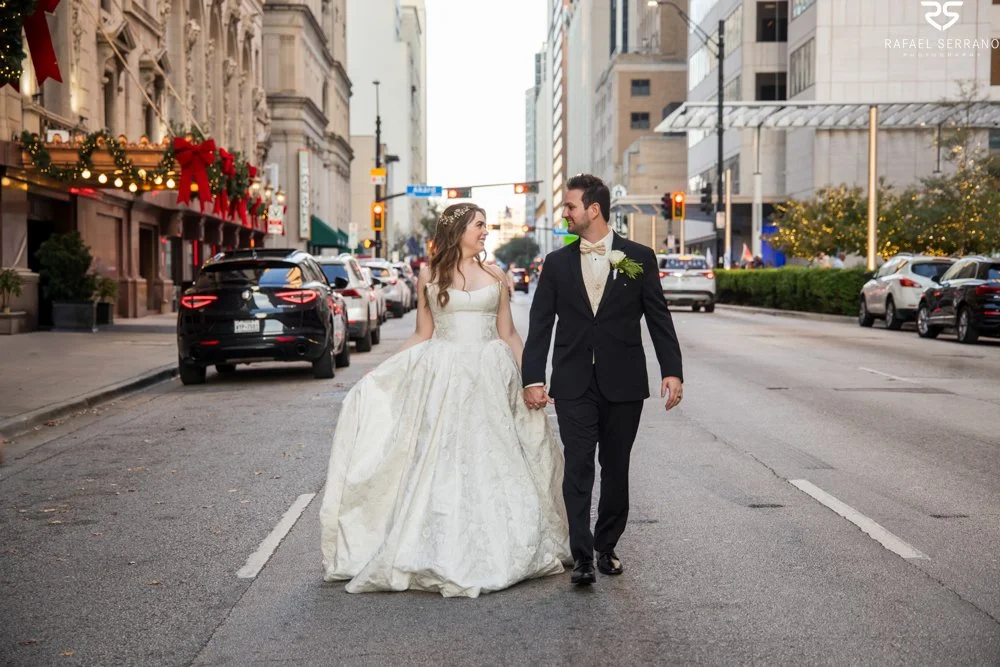 McKenna and Chris walking hand in hand downtown with cars and holiday-decorated buildings behind them.