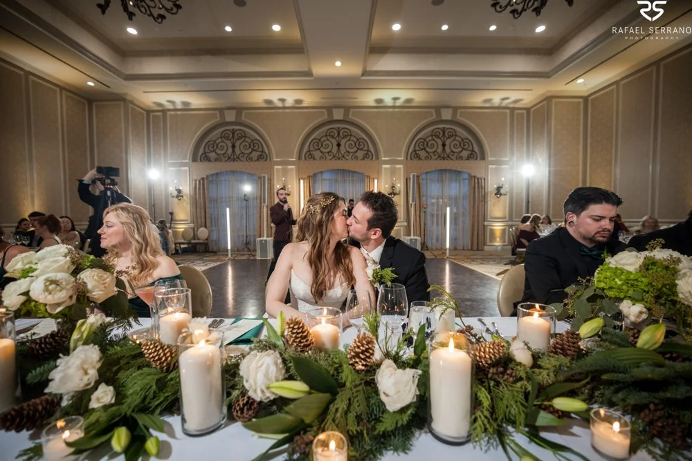 McKenna and Chris kissing at a reception table decorated with white flowers, greenery, and candles.