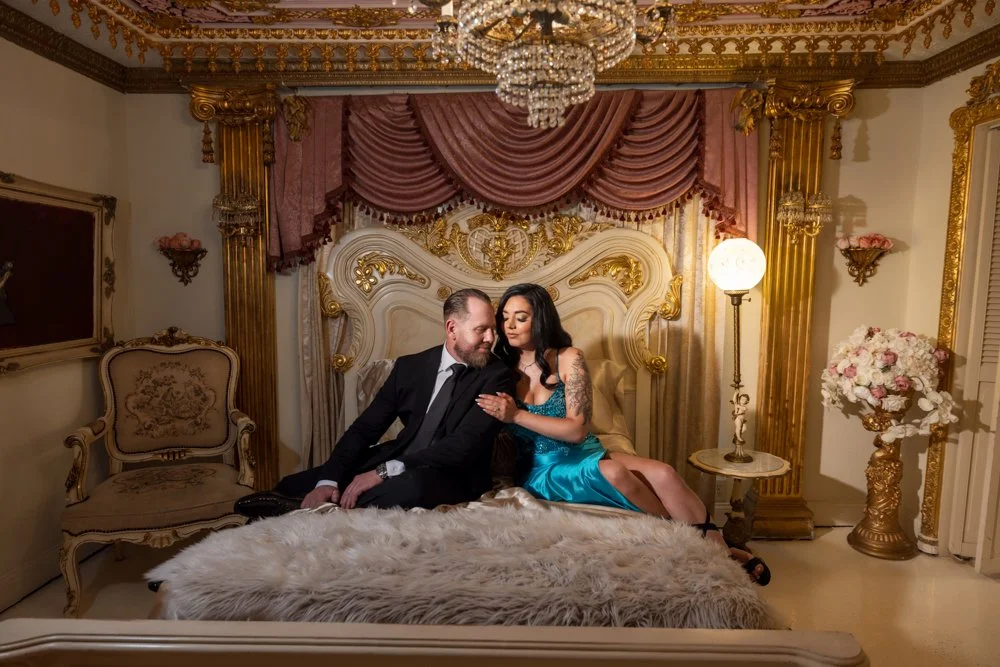 Couple seated together on an ornate bed beneath a chandelier.