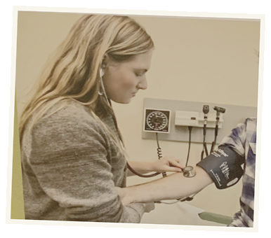 Young woman in a gray sweater taking a person's blood pressure using a sphygmomanometer in a clinical setting.