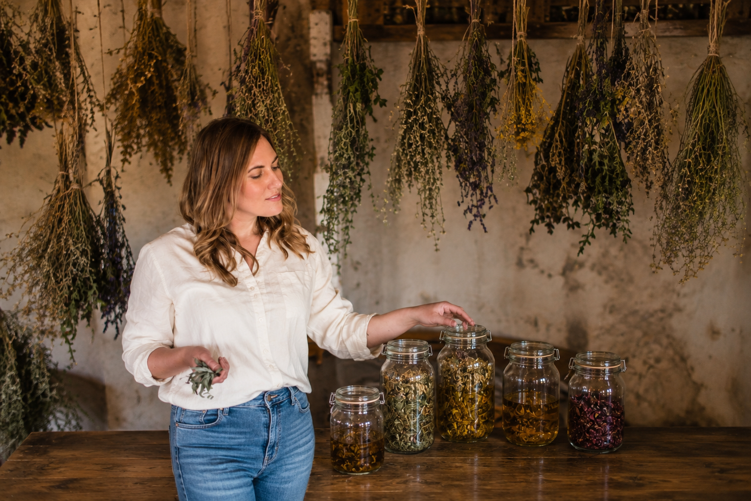 A woman with wavy, shoulder-length hair wearing a white shirt and blue jeans stands at a wooden table. She is holding a small branch in her left hand and opening a large jar with her right hand. On the table are five glass jars filled with various dried herbs or flowers. Behind her, dried herbs hang from the ceiling against a rustic wall.