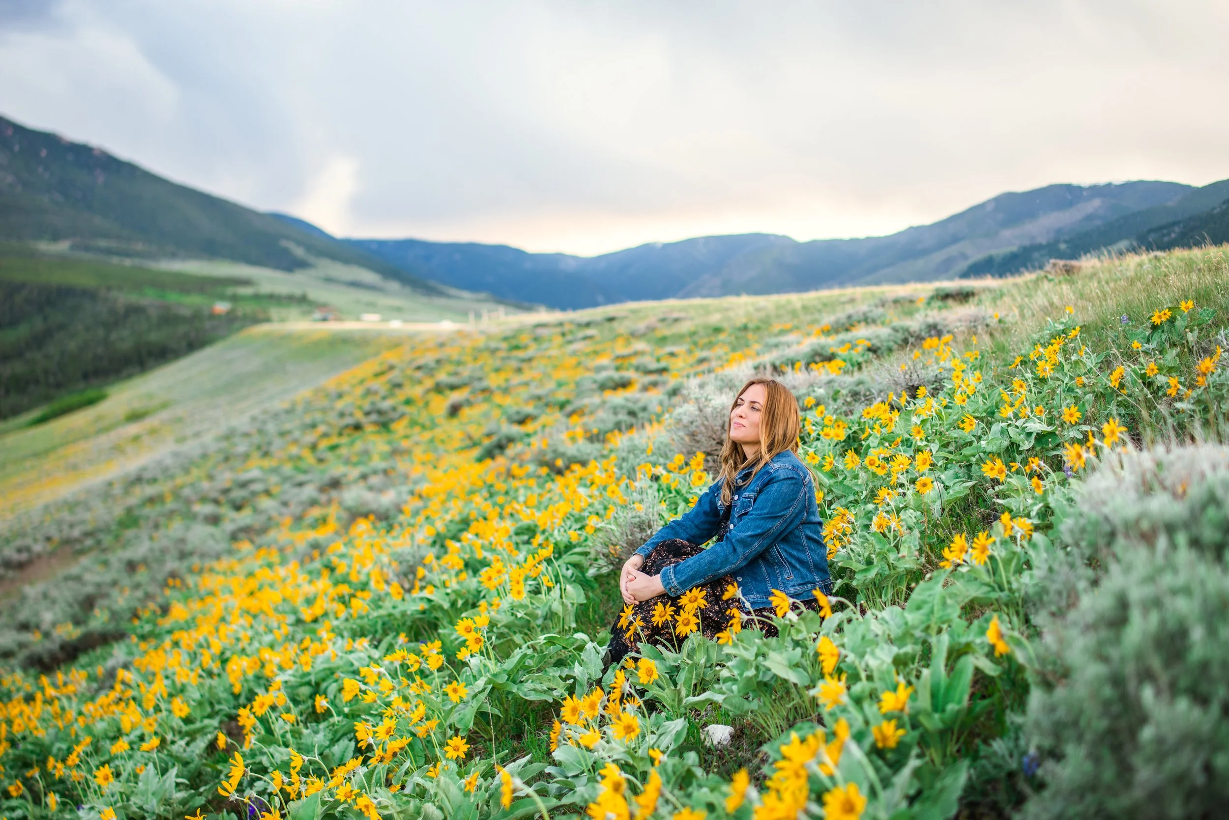 A woman with long hair sitting among yellow flowers in a green field with mountains and cloudy sky in the background.