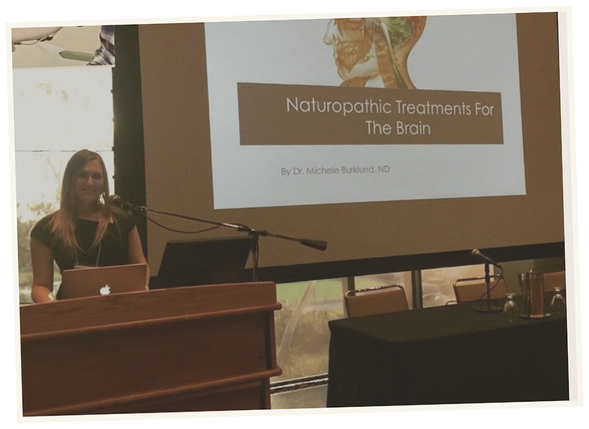 A woman standing at a podium with a laptop, giving a presentation on 'Naturopathic Treatments For The Brain' by Dr. Michele Burklund, ND, with a projected slide showing a brain illustration.