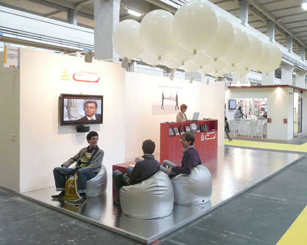 Lo stand con la zona relax in primo piano e il desk sullo sfondo, messo in risalto dai palloni sospesi.