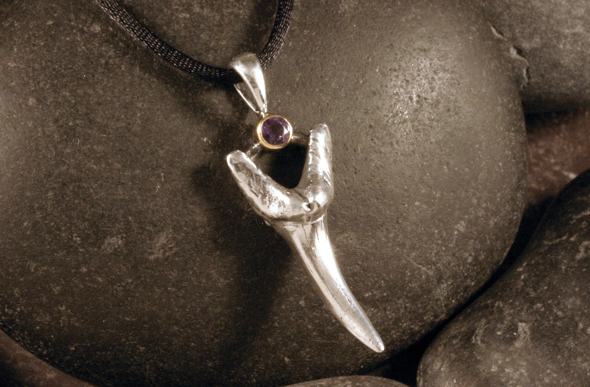 An angled view of a sterling silver fossil shark tooth pendant with an amethyst accent in a 14K gold bezel.