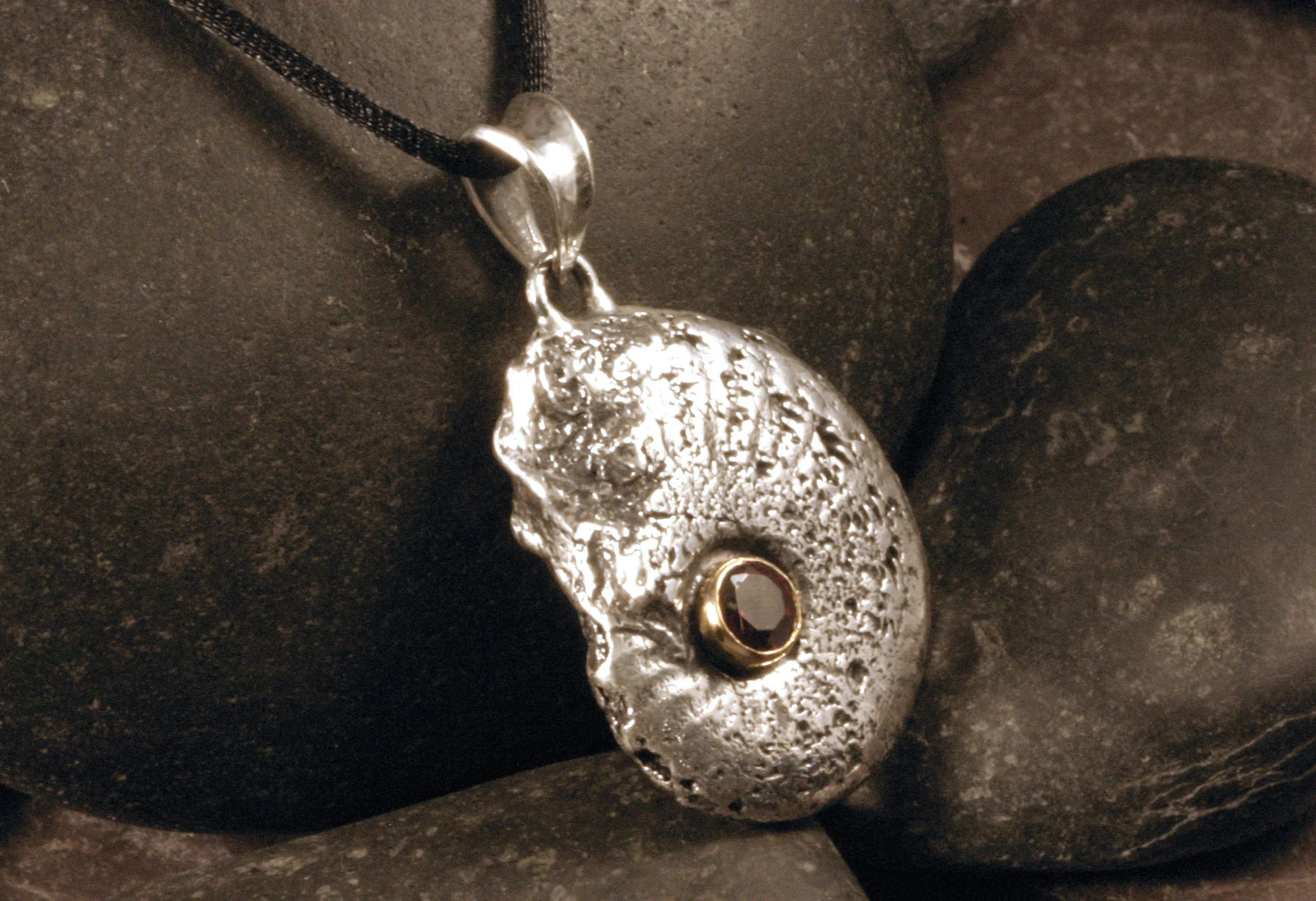 An angled view of a sterling silver fossil ammonite pendant with a red garnet in a 14K gold bezel at the center of its spiral.