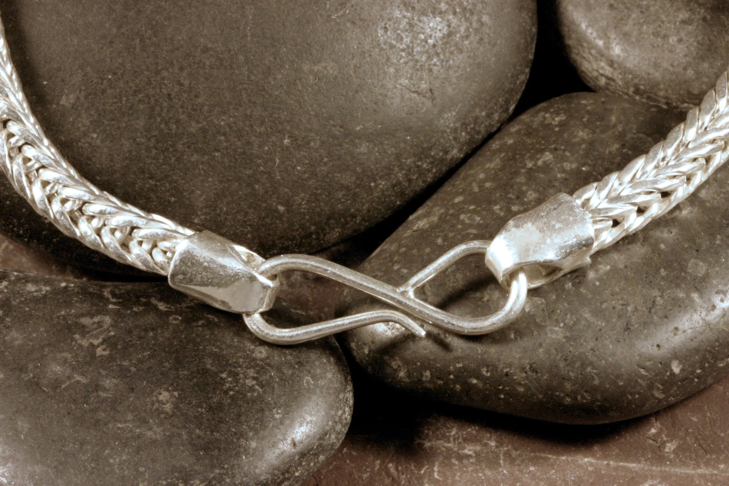A close up of the smooth end caps and S hook clasp on a handwoven sterling silver square double loop in loop chain necklace.