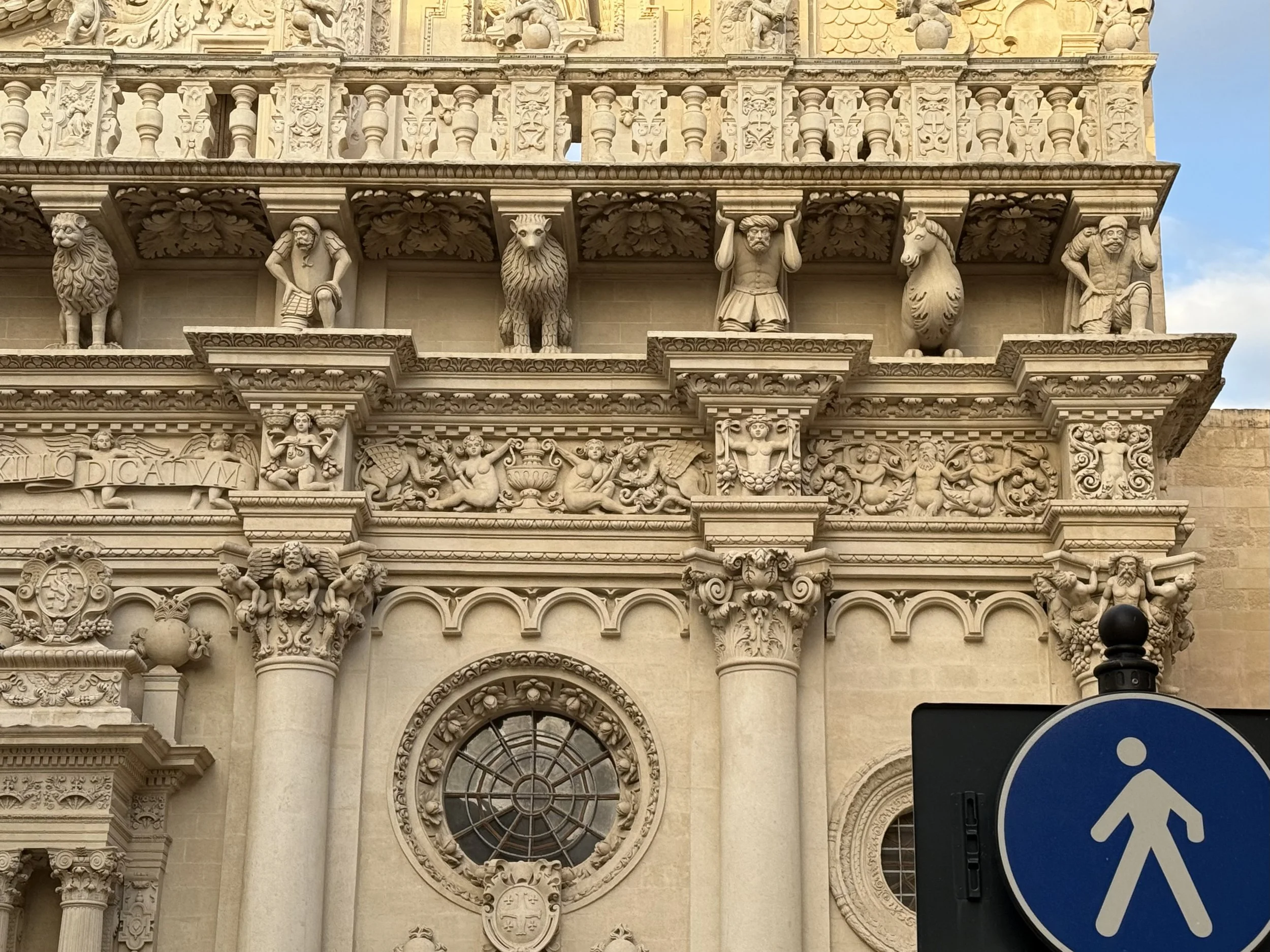 Intricate stone carvings and sculptures on the facade of a historic building, featuring human figures, animals, and decorative elements, with a large arched window at the center.
