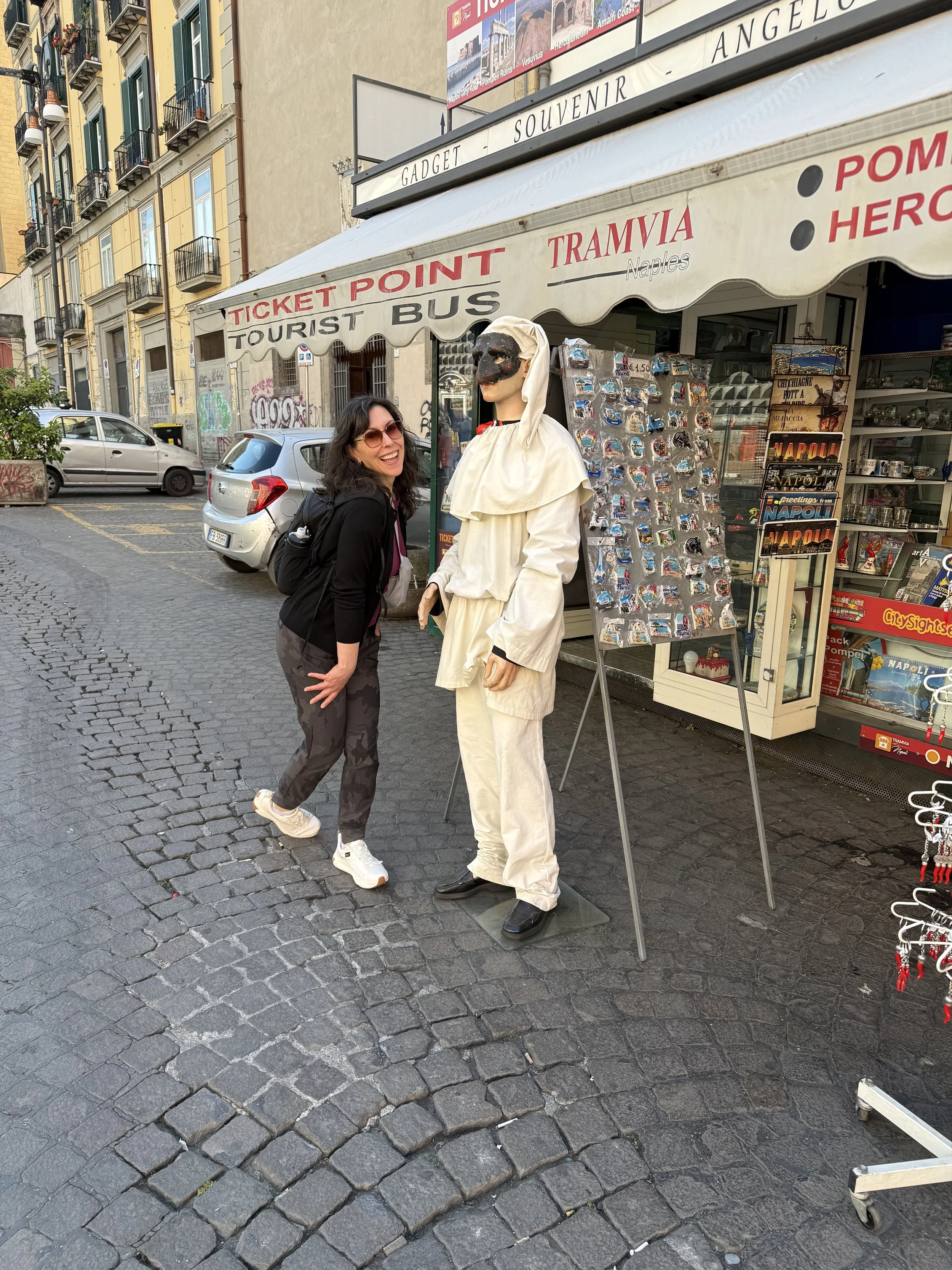 A woman with dark hair and sunglasses, smiling and holding her knees, standing next to a mannequin dressed as a traditional person in a white costume with a black mask and hood, outside a souvenir shop on a cobblestone street.