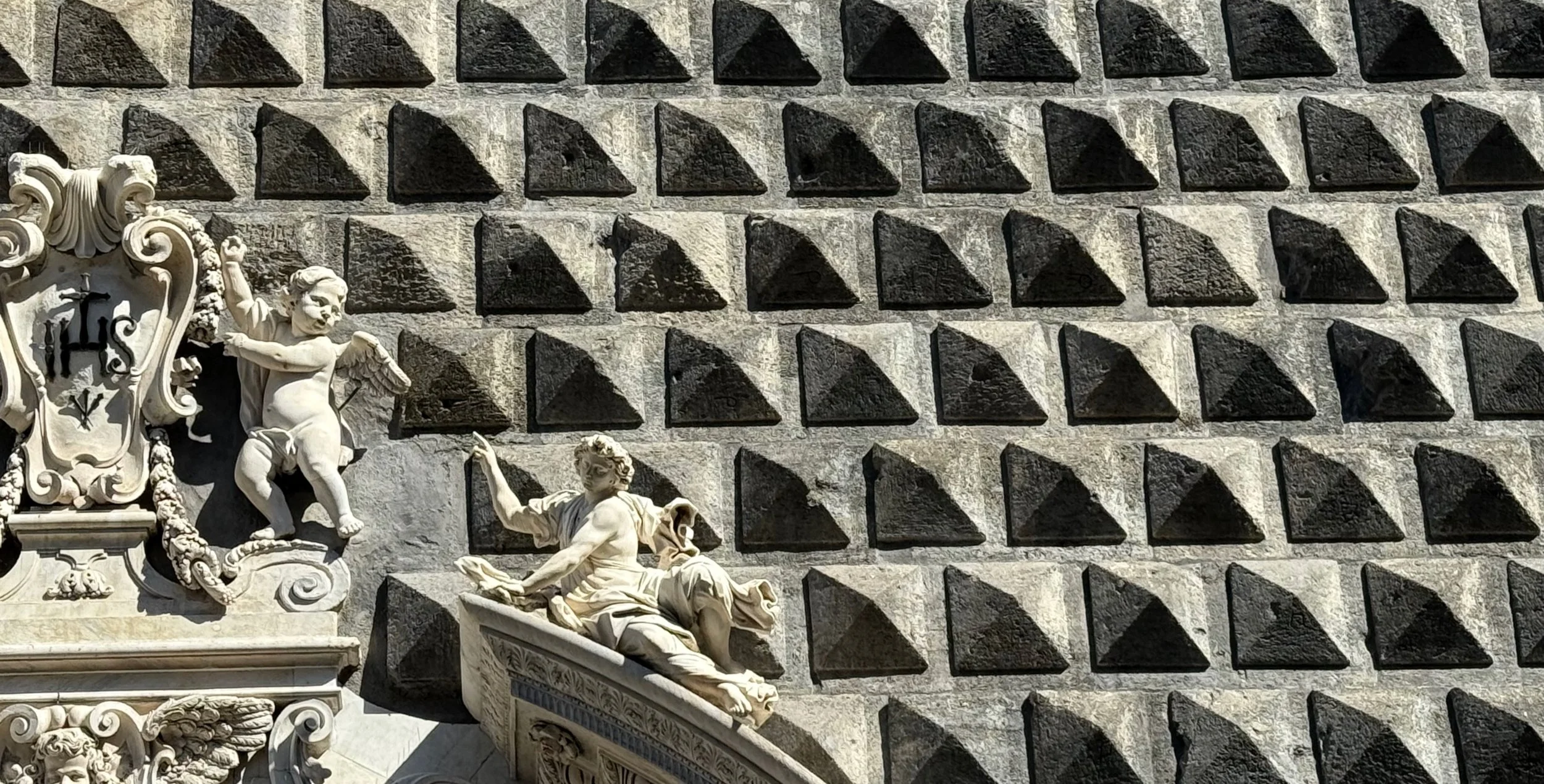 Close-up of a decorative stone wall with geometric pyramid-shaped details and classical sculptures of figures and a shield.