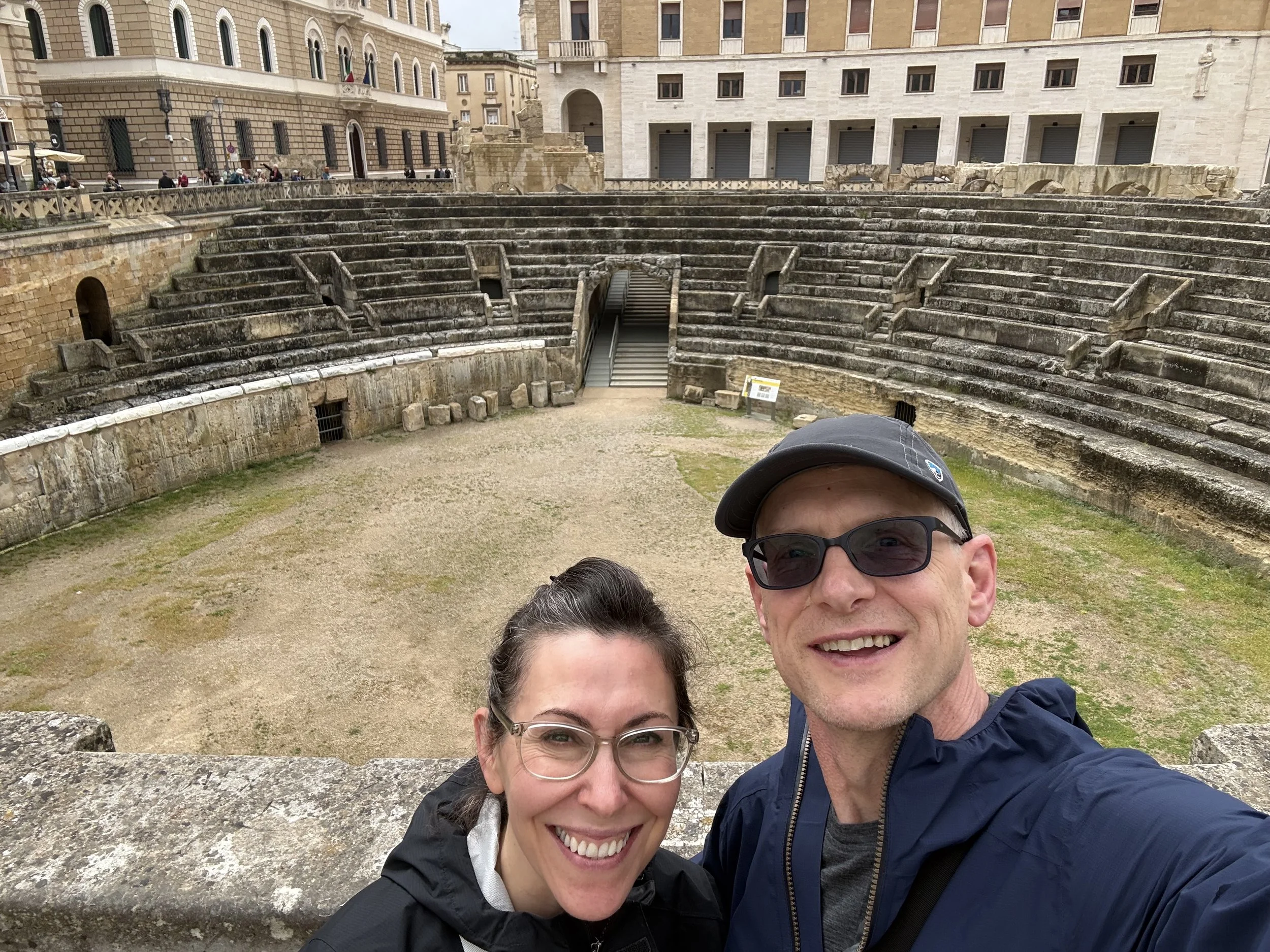 A group of three smiling people taking a selfie in front of ancient Roman ruins, with a partially preserved amphitheater and historic buildings behind them.