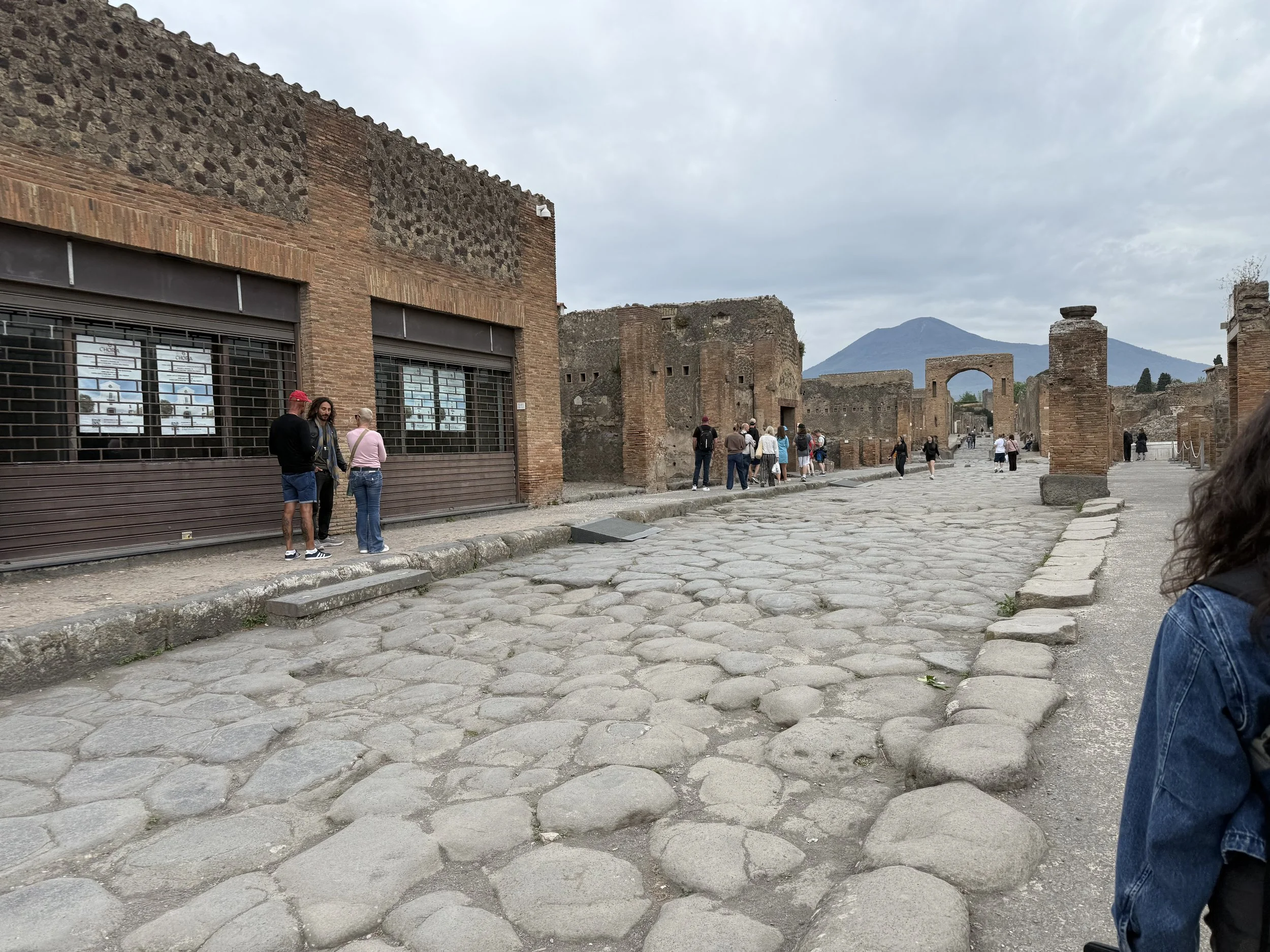 Tourists walking along ancient ruins with stone pavement and brick walls, with mountains in the background under an overcast sky.
