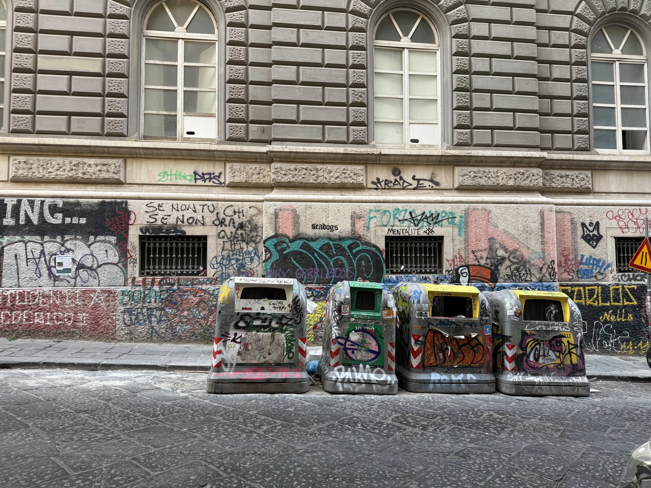 Street scene with graffiti-covered building, graffiti-covered trash bins, and parked cars.