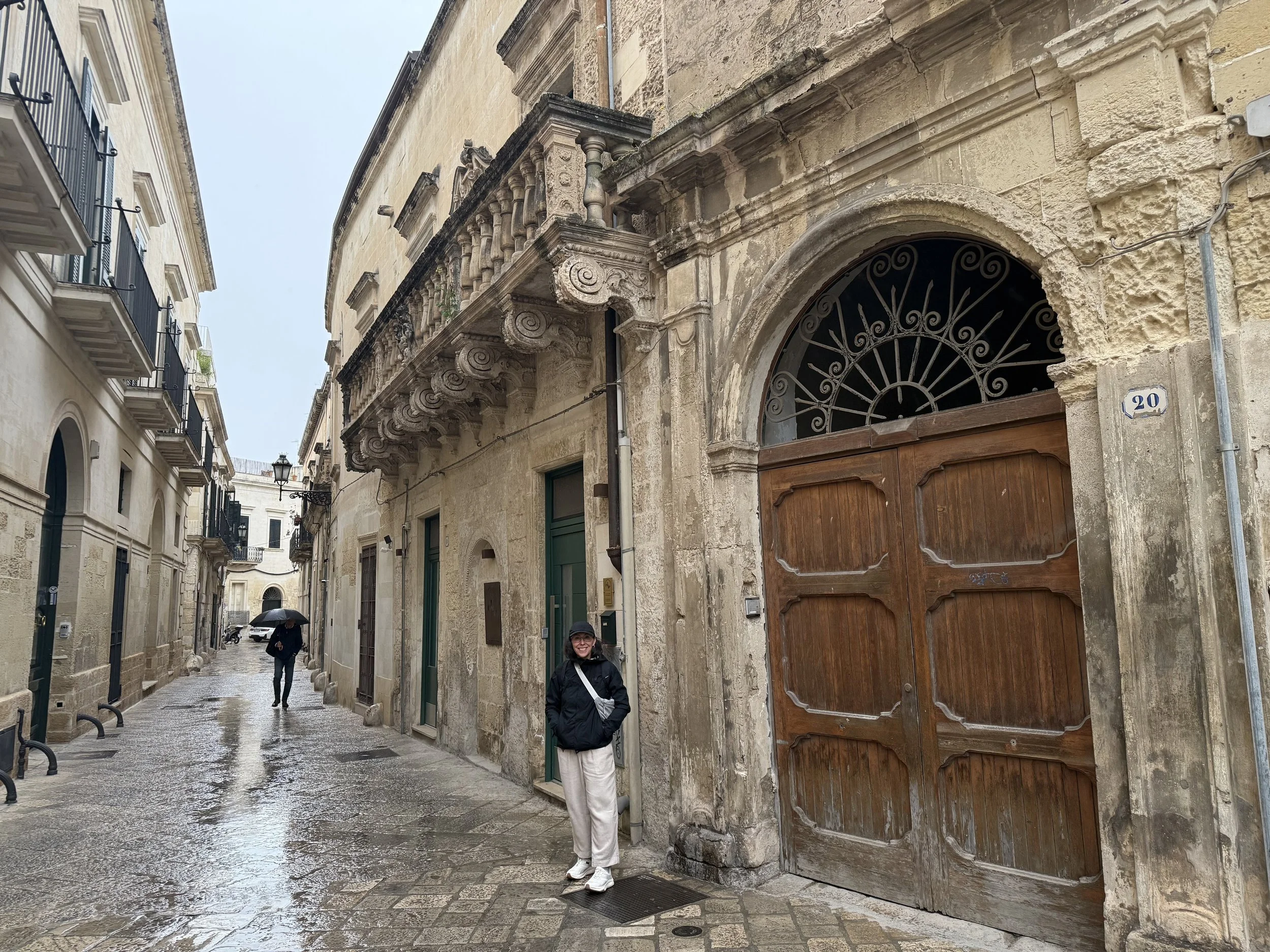 A woman standing in front of large wooden doors on an old stone building. The street is wet, and there are two people walking behind her, one holding an umbrella. The architecture has ornate detailing and balconies.