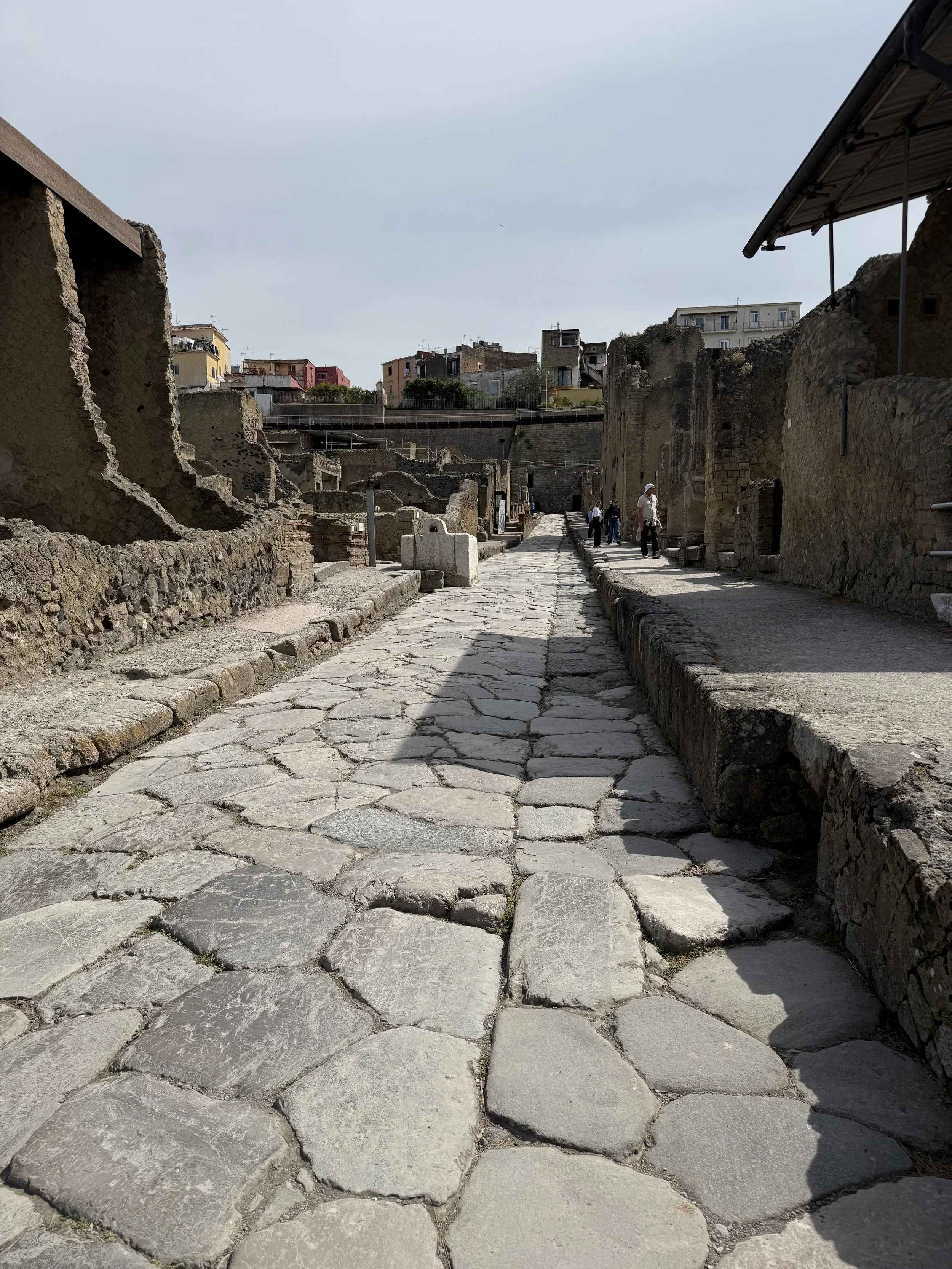 Ancient stone street with ruins, modern buildings in the background, and a few visitors exploring the site.
