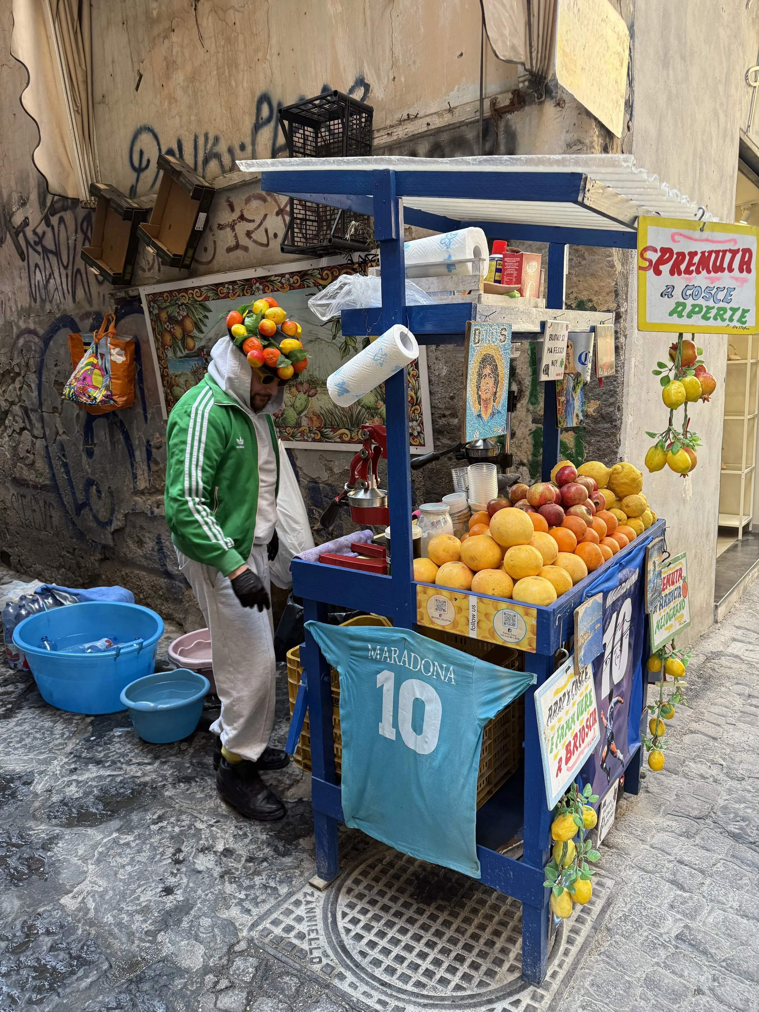 A man in a green jacket with cucumbers on his head working at a fruit stand, selling various fruits including oranges, lemons, and apples, on a cobblestone street.