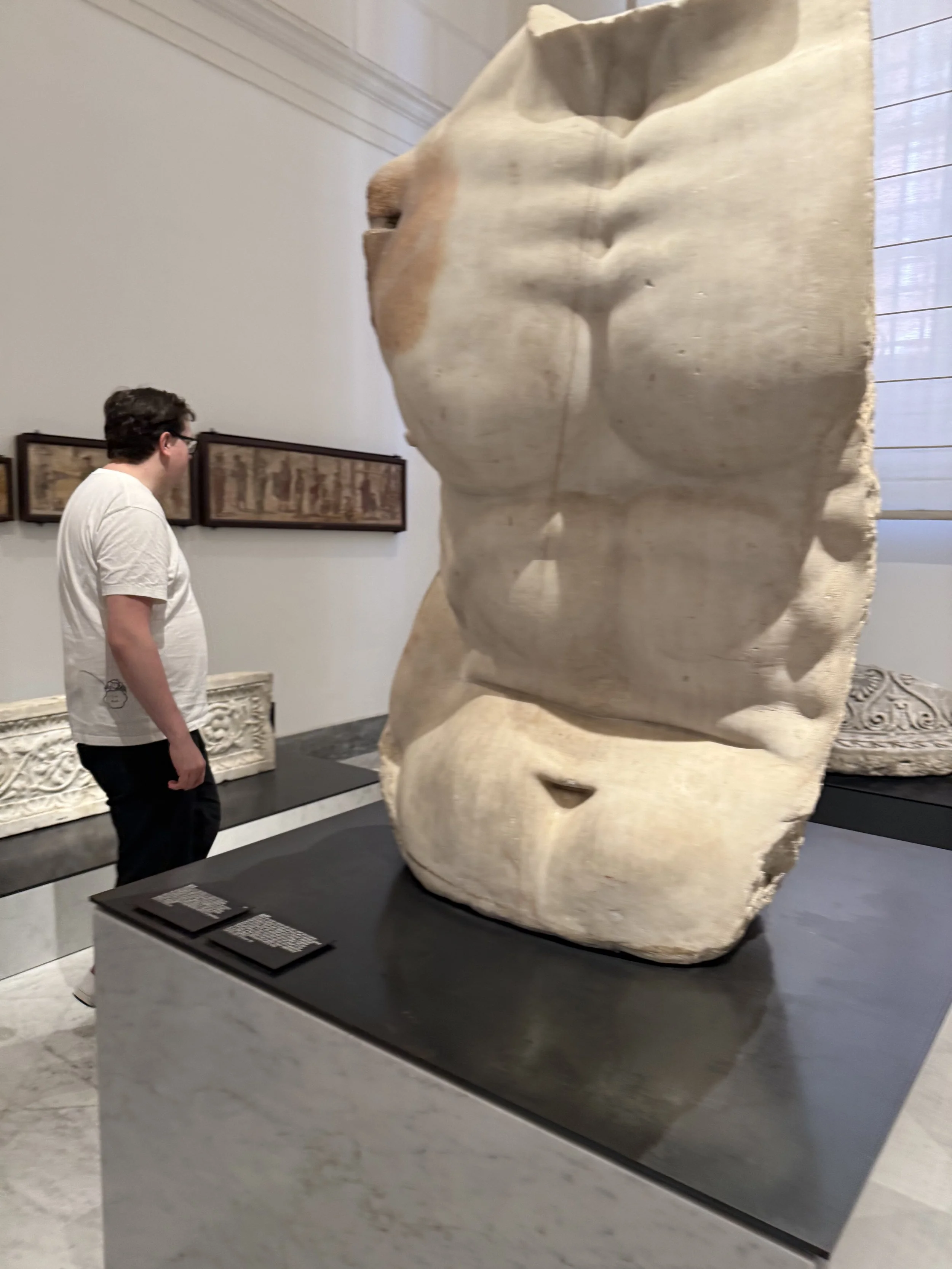 A person standing in a museum looking at a large ancient stone sculpture of a human face with closed eyes.