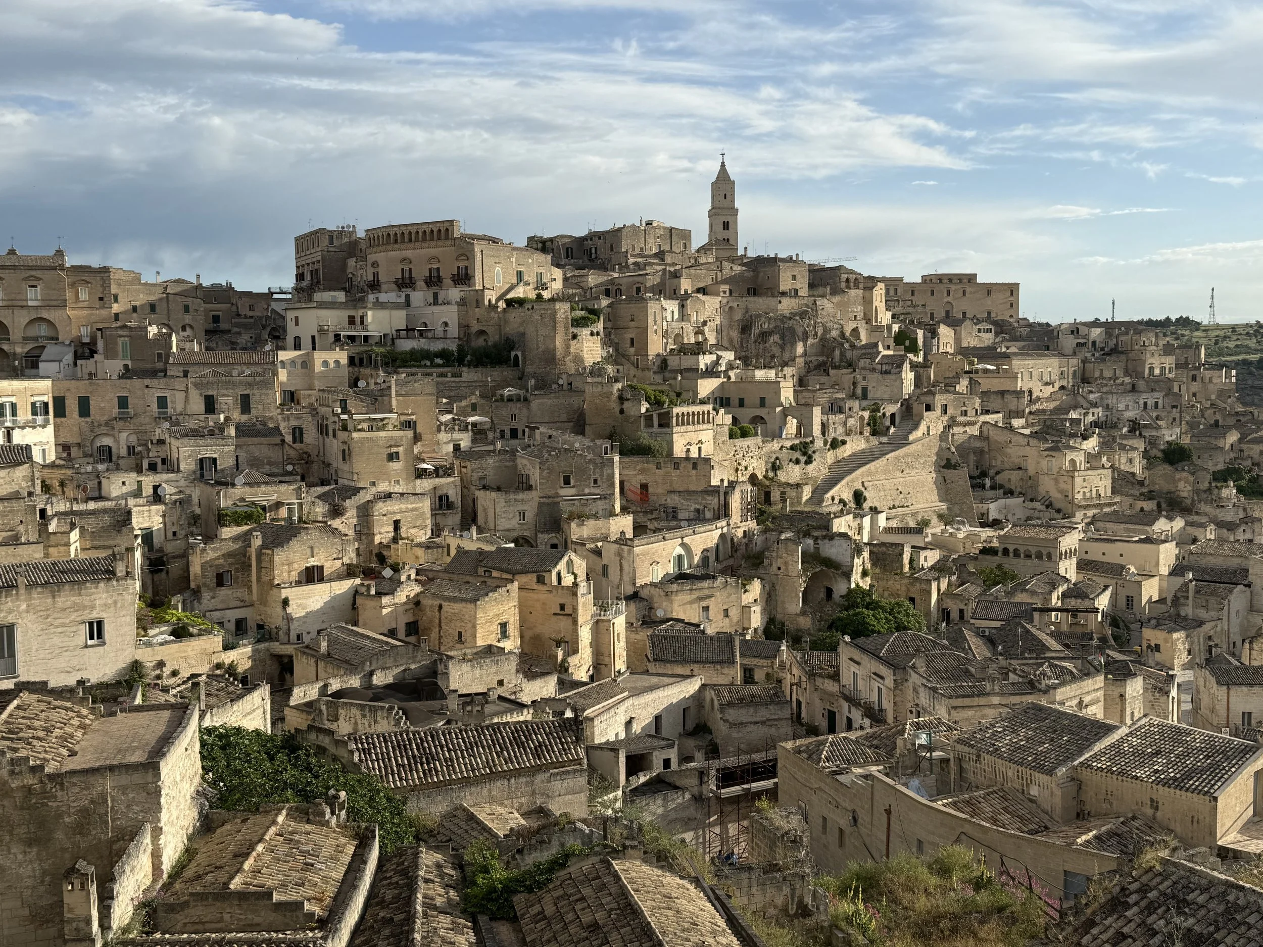 A historic city with stone buildings and narrow streets, with a tower in the background under a partly cloudy sky.