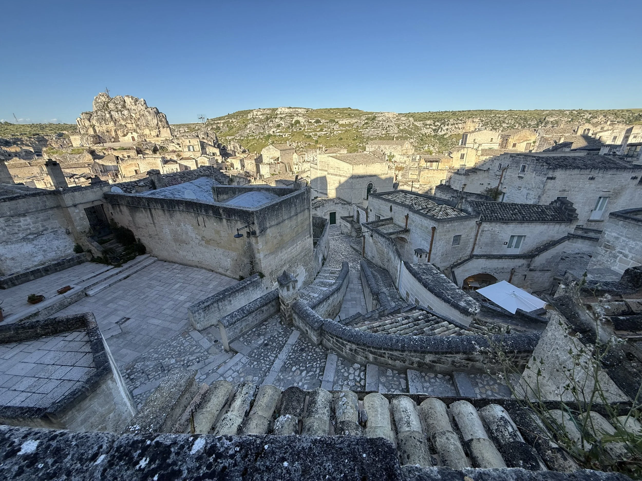 A stone staircase with railings leads down a historic stone city with multiple rooftops, surrounded by rocky hills under a clear blue sky.