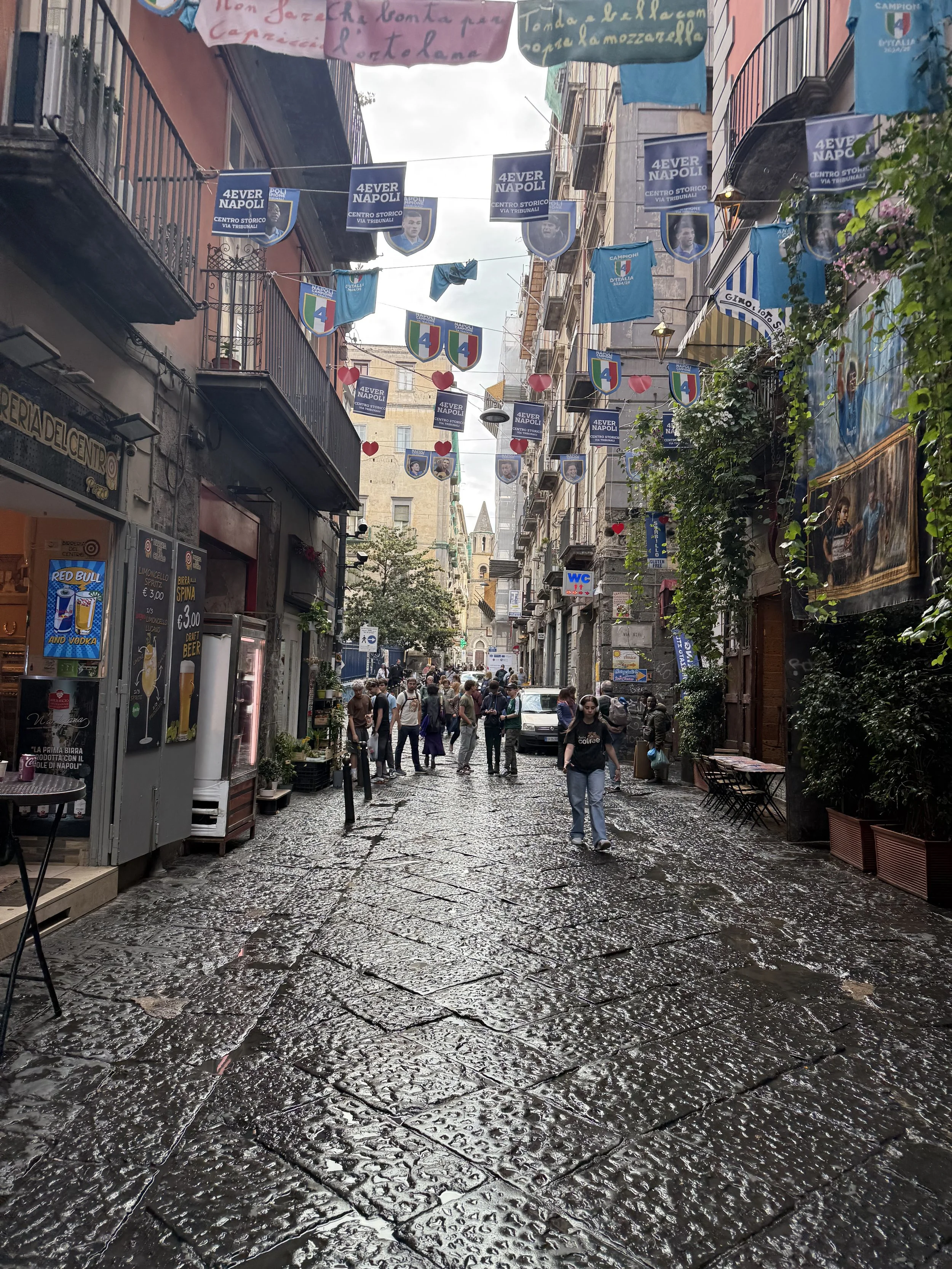 A narrow city street with wet cobblestone pavement, lined with cafes and shops, decorated with flags and banners, and filled with pedestrians.