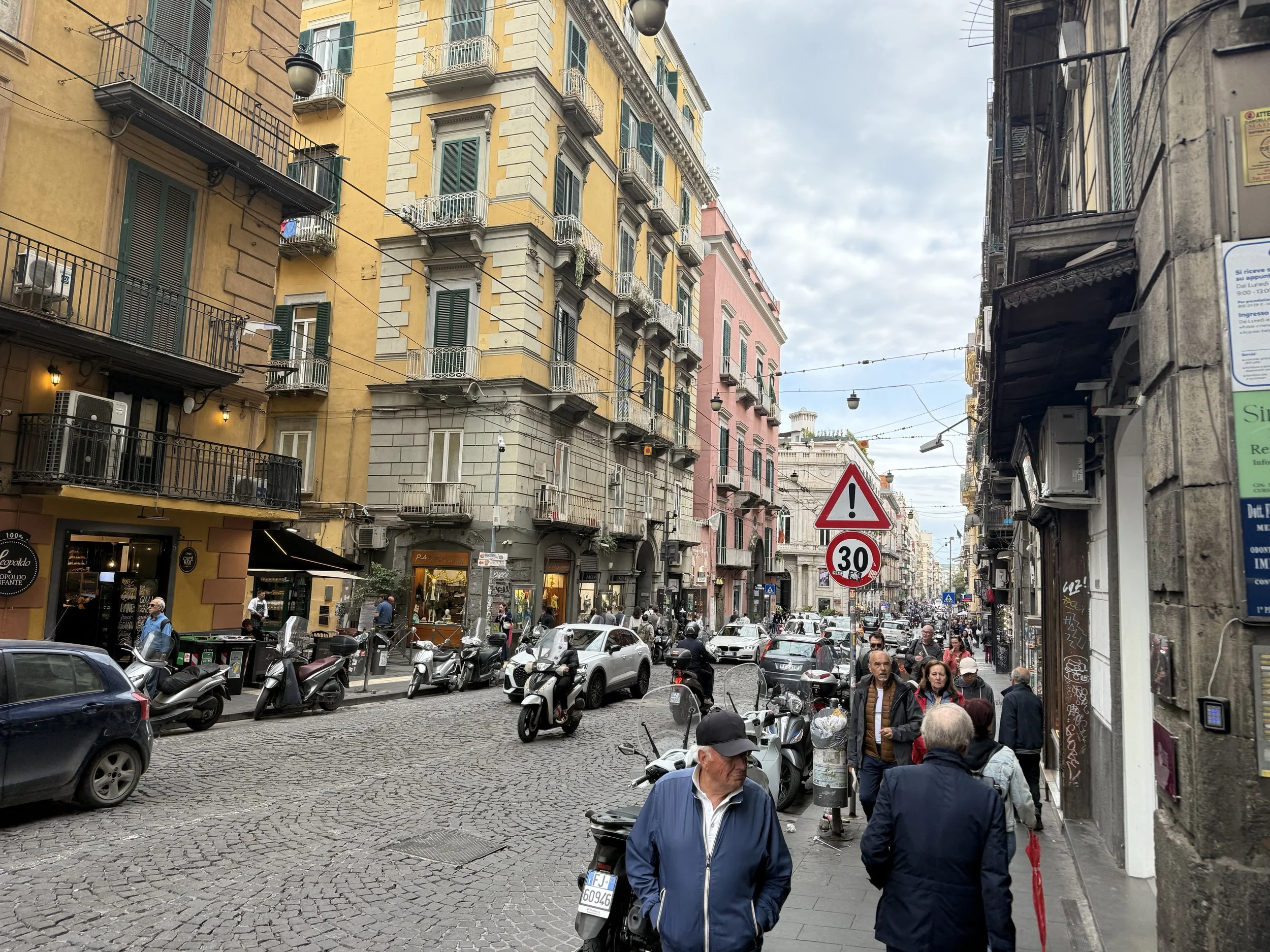 A busy city street crowded with pedestrians and parked scooters. Multi-story colorful buildings line the street, with some people walking on the sidewalk and cars visible on the road. The sky is partly cloudy.