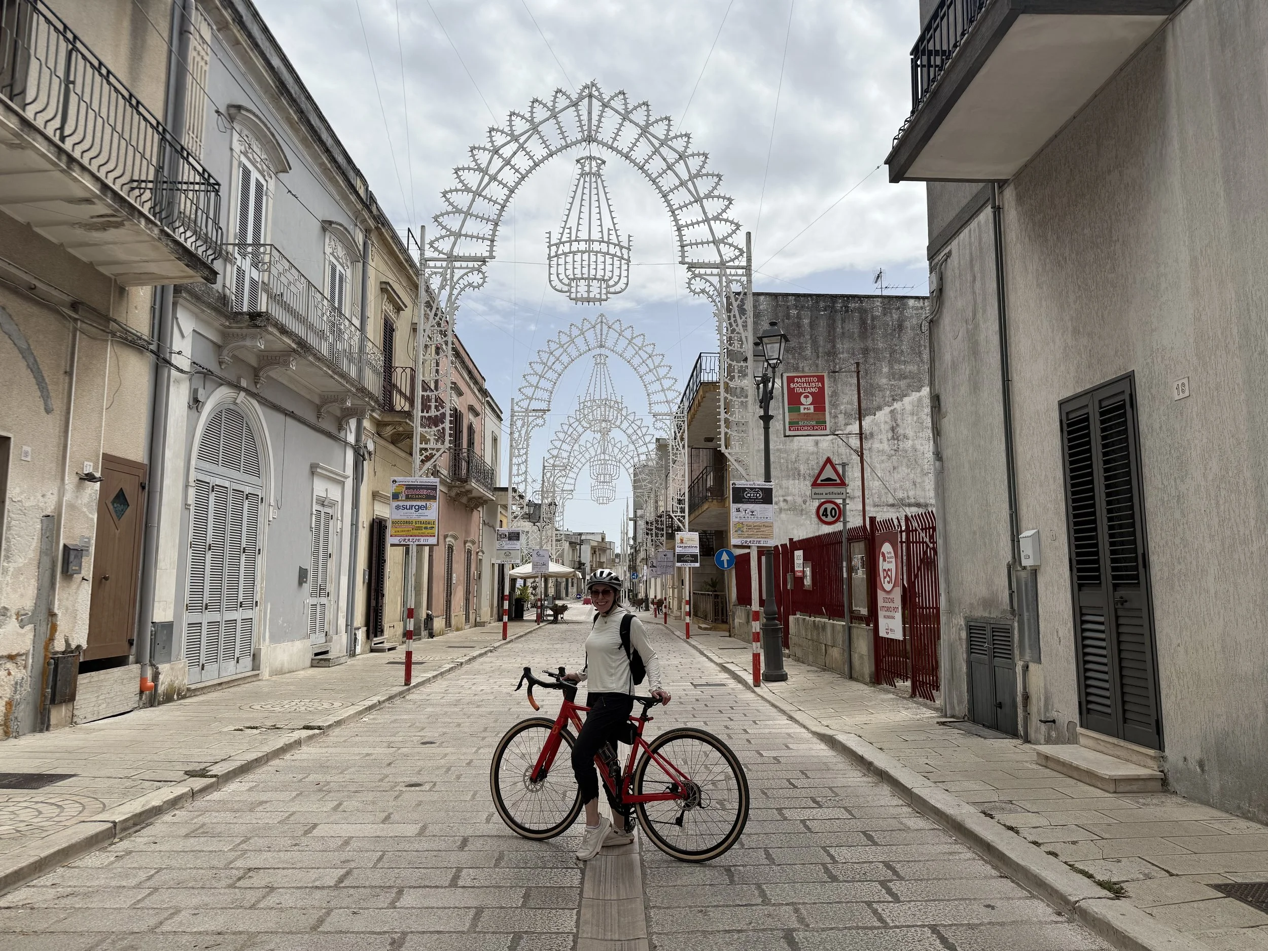 A woman with a bicycle standing on a cobblestone street in a European city with decorative street lights overhead, surrounded by old buildings with balconies and shutters.