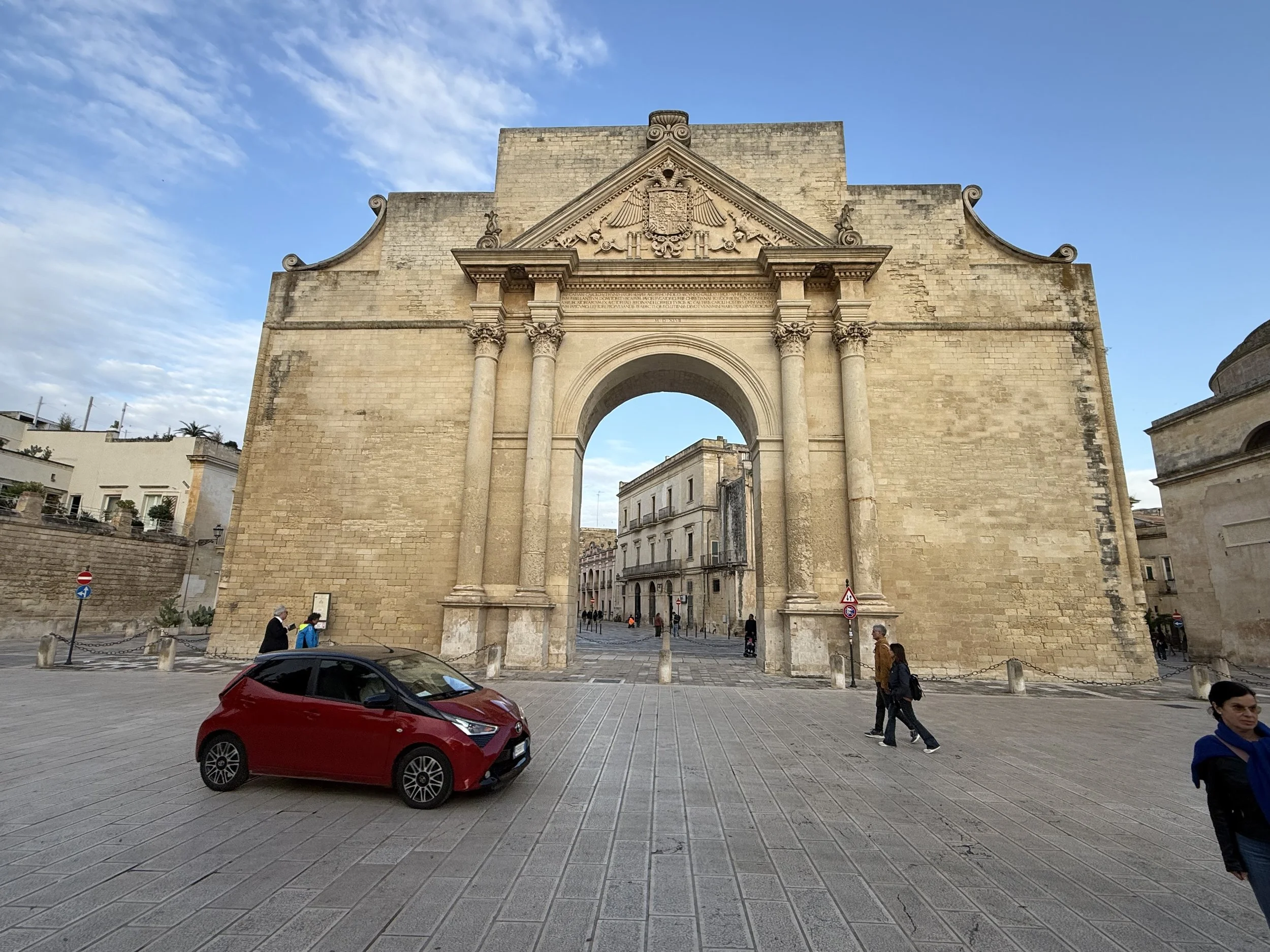A historic stone archway with ornate carvings and a coat of arms at the top, situated in an urban area with people walking and a red car parked in the foreground.