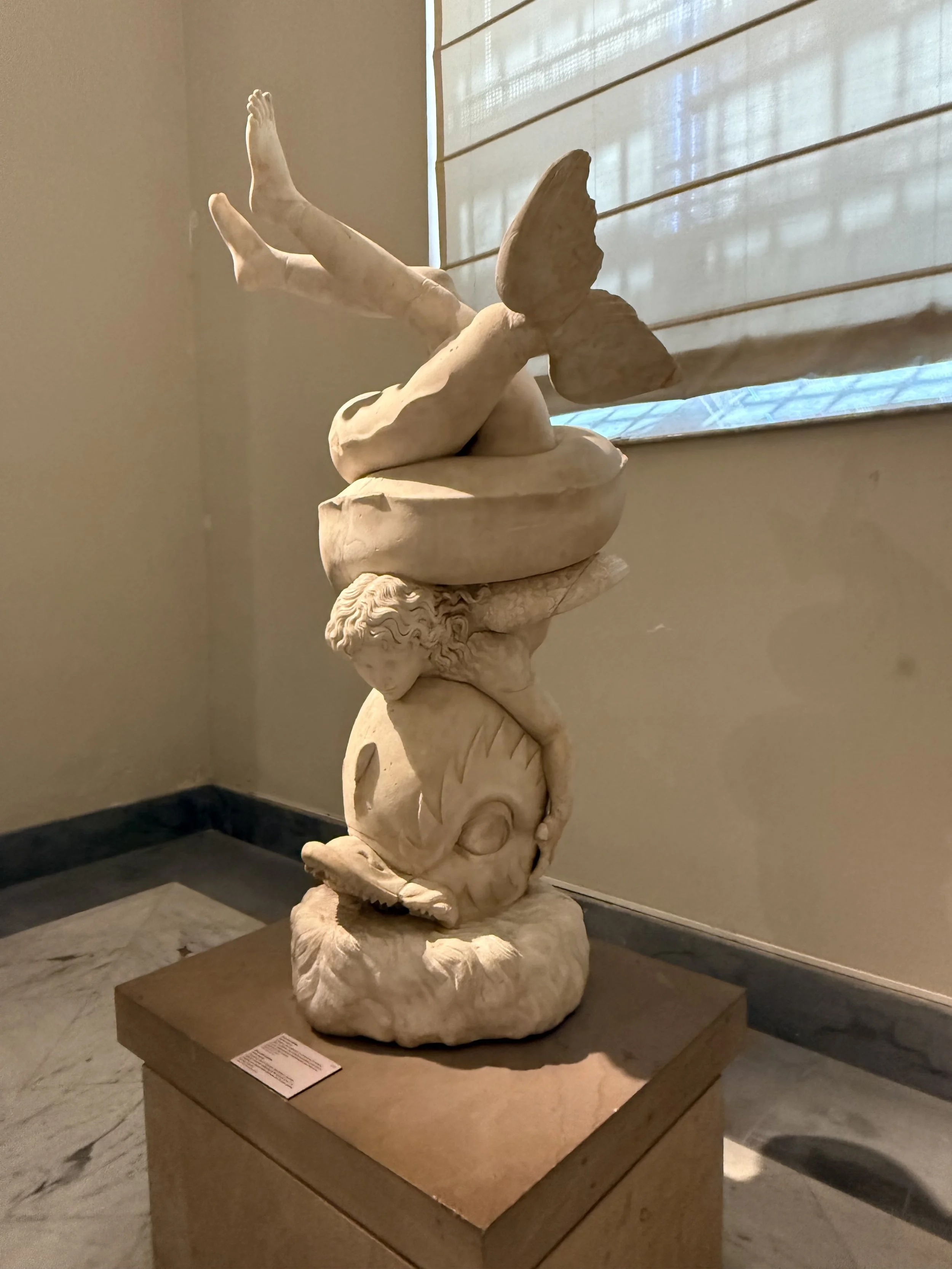 A marble sculpture of a reclining female figure with wings, lying on a base that resembles a fish, displayed on a wooden pedestal indoors near a window with blinds.